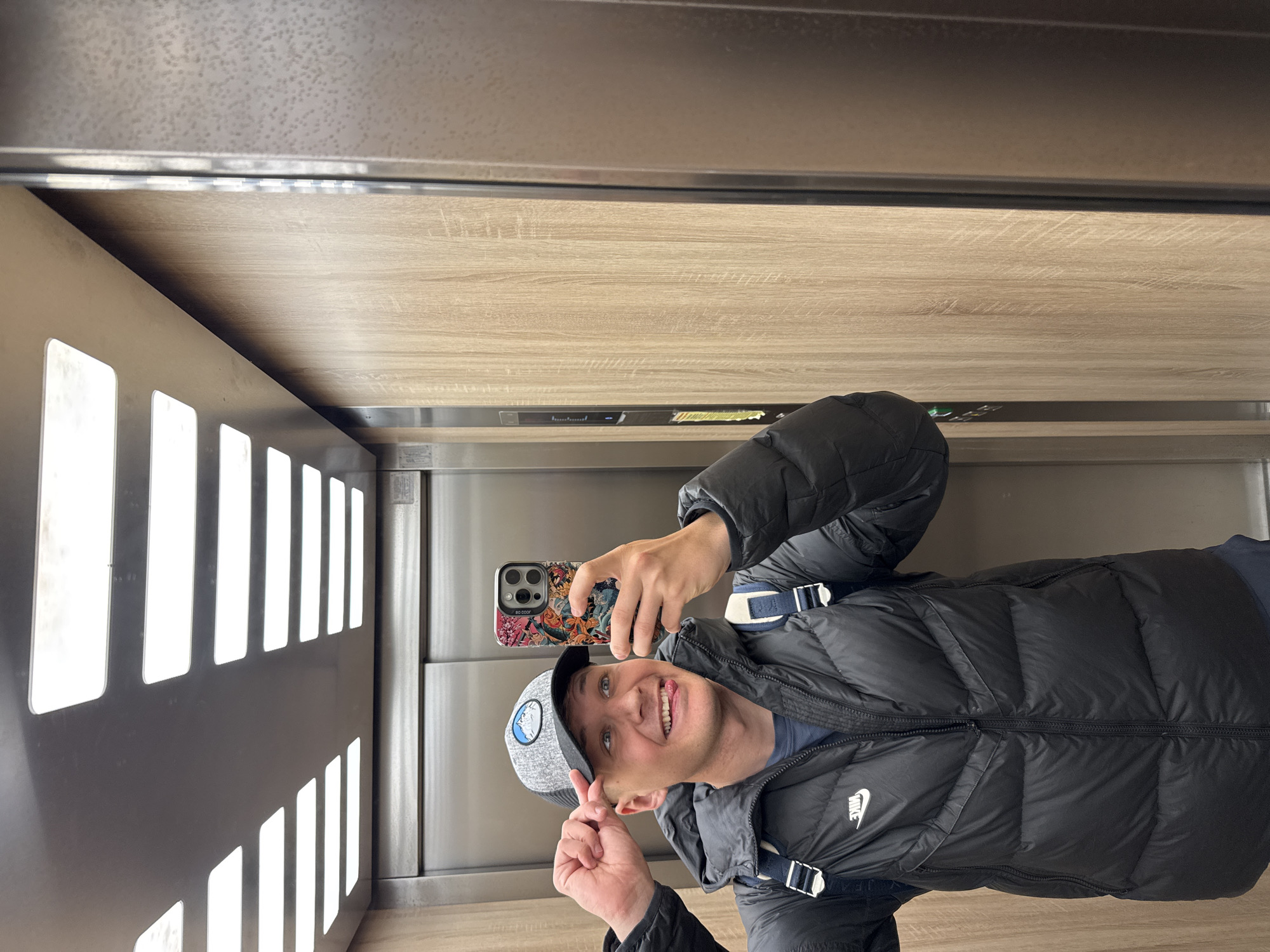 The image depicts a man taking a selfie in what appears to be an elevator. He is wearing a black puffy jacket, a grey baseball cap with a logo, and holding a smartphone with a patterned case. He is smiling at the camera and holding up two fingers in a peace sign gesture. The elevator interior consists of wood-paneled walls and a metal panel with rectangular cutouts. The location is an elevator in Plovdiv, Bulgaria.