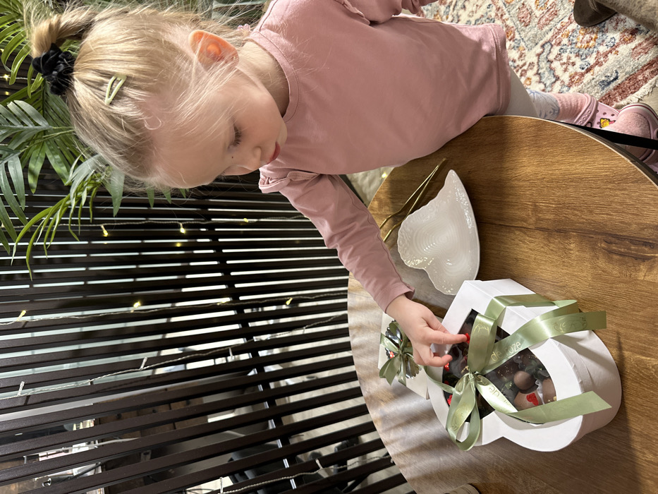 A young child with blonde hair in a ponytail, wearing a pink shirt, reaches for a chocolate-filled, heart-shaped box on a round wooden table. A heart-shaped plate sits nearby. The background features a decorative screen with horizontal slats and fairy lights, along with a potted plant. The floor has a patterned rug. The overall scene suggests a sweet, festive occasion.