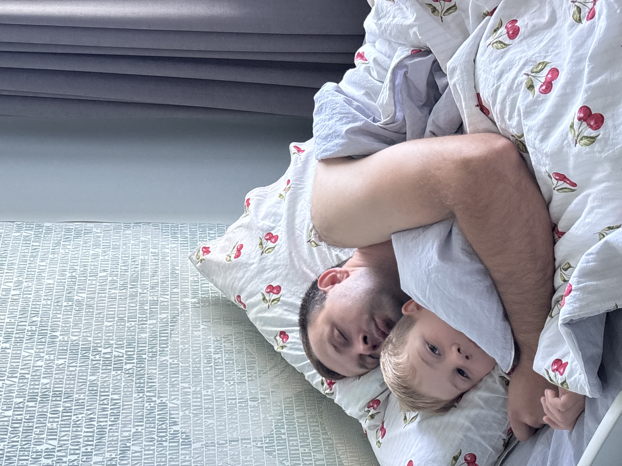 In Rusanovo, Russia, a father and son are asleep in bed. The father is lying on his back, while the son is nestled in his arms, facing the camera. Both are under a white duvet patterned with red cherries. The background consists of a wall with text and grey curtains. The lighting is soft, indicating natural light from a window.