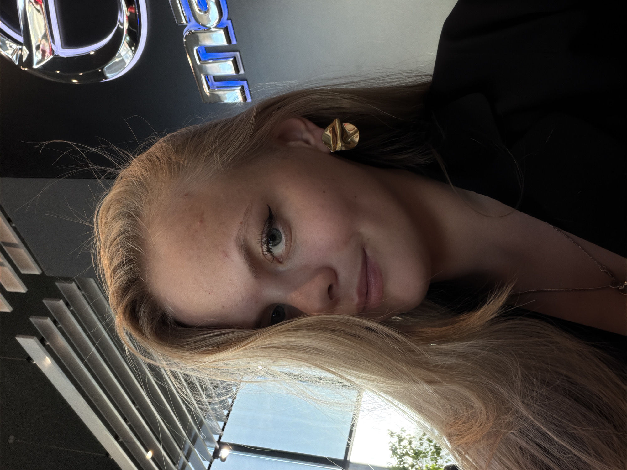 A woman with long blonde hair and fair skin is facing the camera. She is wearing a black blazer. Her left ear is adorned with a gold, crumpled-looking earring. Behind her is a sign with reflective chrome lettering outlined in blue. In the bottom portion of the image, a window reflects the sky and trees. The setting appears to be indoors, possibly a retail or commercial space, as indicated by the sign and the louvered ceiling fixture in the lower-left corner. Without additional information, determining the precise location in Izhevsk, Russia, is not possible.