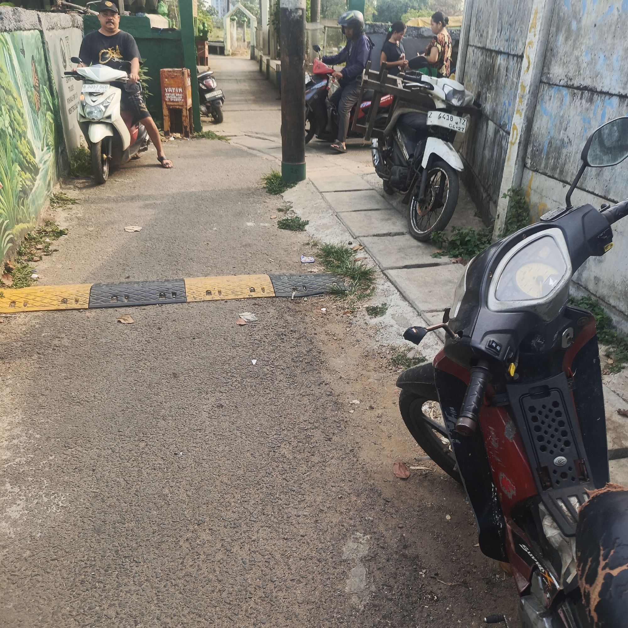 The image depicts a street scene in South Jakarta, Indonesia, characterized by an asphalt road with a speed bump in the foreground. On the left side of the road, a man wearing a black t-shirt, shorts, and a cap sits on a white scooter. He is positioned next to a wall adorned with a mural of greenery and a butterfly. Further down the street, two women are standing near a wall with another scooter parked beside them. Several other motorcycles are visible parked along the street. The environment appears to be an urban residential or commercial area, with buildings and vegetation lining the walkway. The lighting suggests it is daytime, with shadows indicating the sun's position.