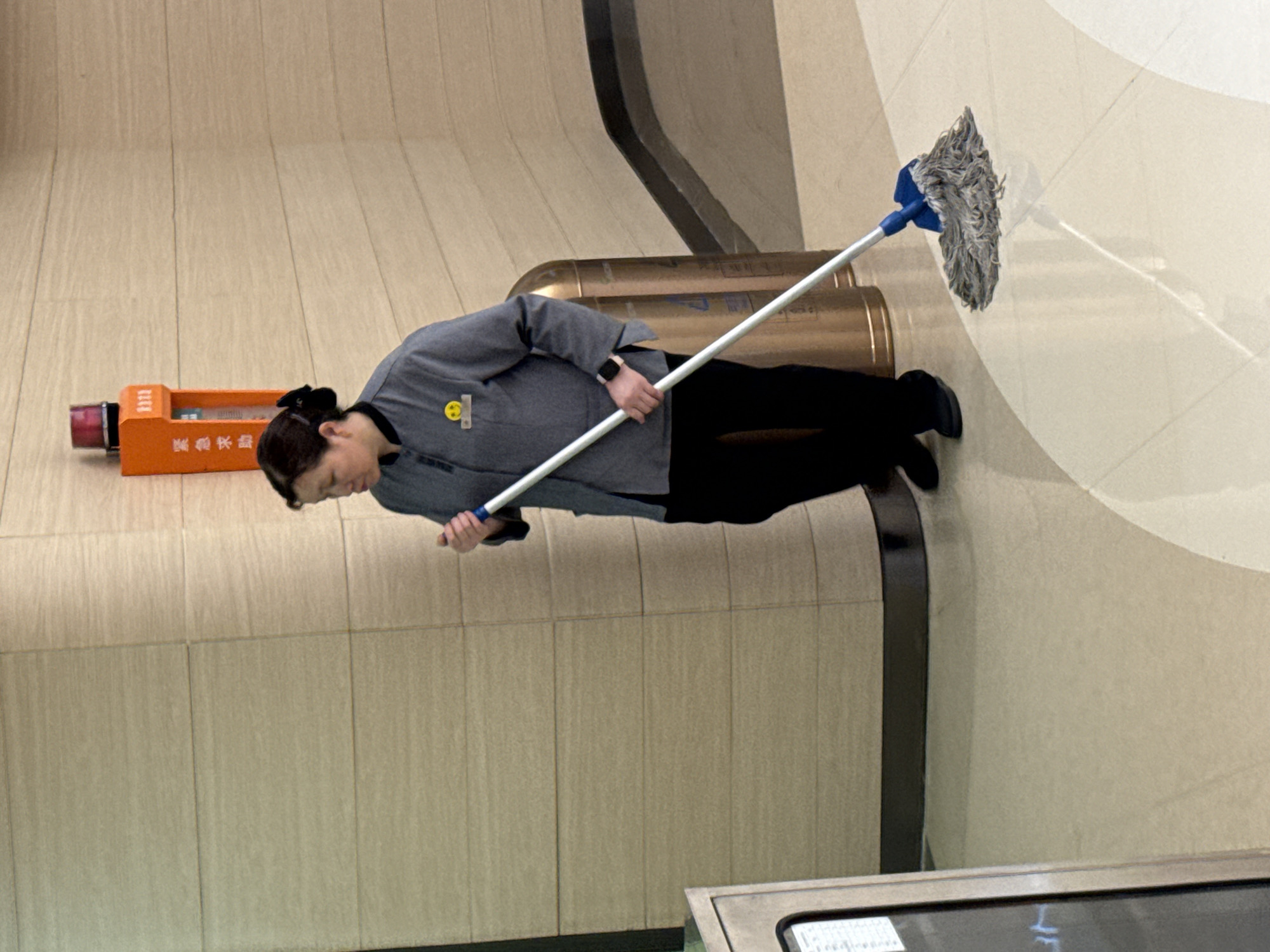 A woman in a grey uniform and black pants is mopping a beige tiled floor. She is holding a mop with a blue handle and a grey, stringy mop head. To her left is an orange object, and behind her are cylindrical objects. The environment appears to be an interior space. Based on the building signage, the location is in Liangjiang, China.
