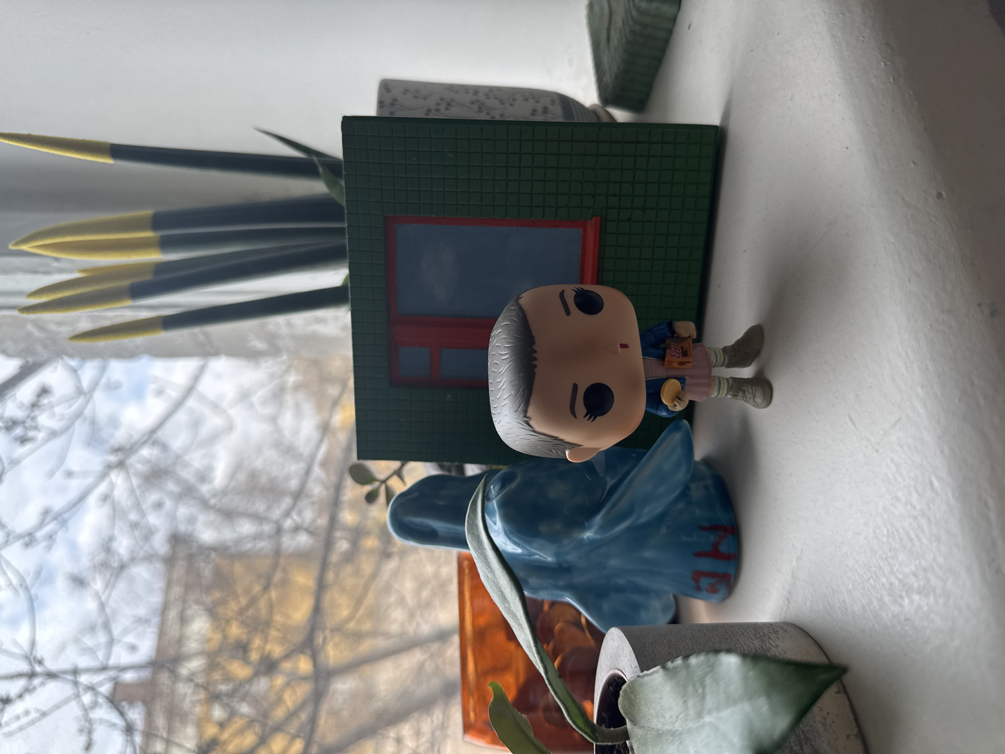 The image shows a close-up view of a windowsill indoors, with several objects arranged on it. A Funko Pop figure of the character Eleven from the TV show "Stranger Things" is positioned near a green decorative miniature building with a red window frame. Behind these are some tall, slender artificial plants with black stems and yellow tips. To the left, the window allows a view of the outside, showing tree branches and buildings, but the location is not discernibly Moscow, Russia. Below Eleven, there's a blue ceramic figurine with the word "HOME" partially visible in red letters, situated near a small potted plant with green leaves in a gray pot and an orange container. The windowsill is white.