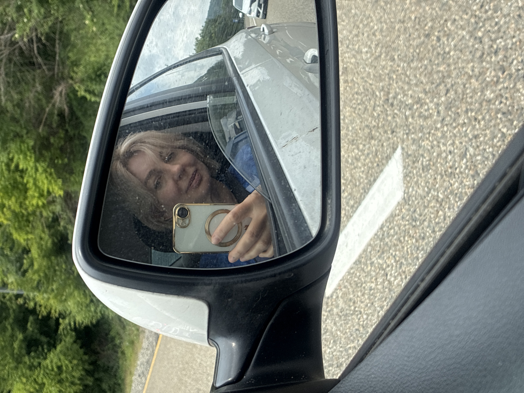 A woman is captured in a reflection on a car's side mirror, taking a selfie with her phone. She has blonde hair and is smiling. The background visible through the car window shows lush green trees and a cloudy sky. The mirror itself is attached to a white car. The reflection also shows a portion of the road with a white line, and another white car is visible further down the road. The environment suggests a highway or a road bordered by nature, consistent with locations like the highway leading to Arkhipo-Osipovka, Russia, which is known for its scenic routes and natural beauty.