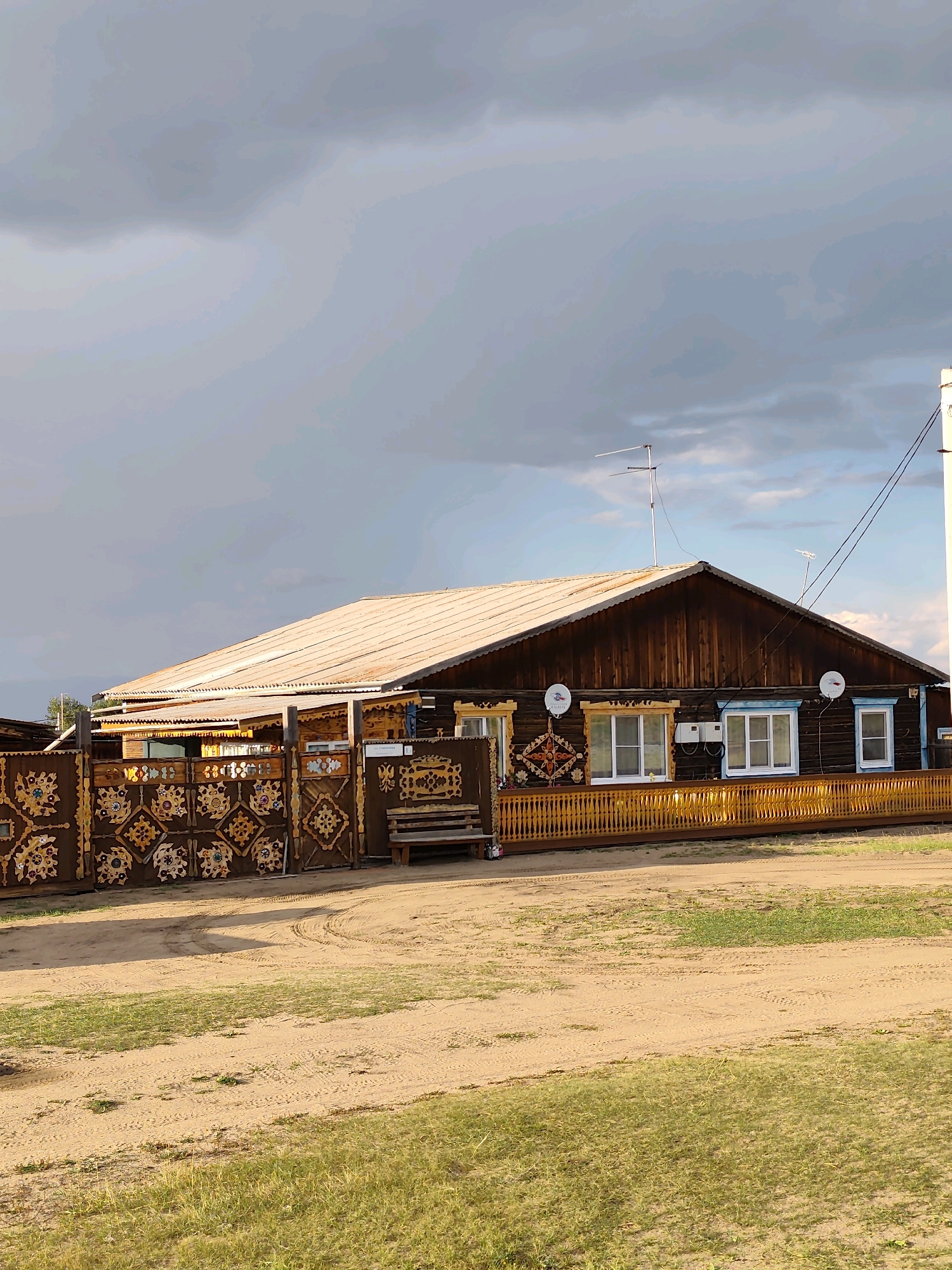 The image depicts a traditional wooden house in the village of Kharantsy, Russia. The house features intricate brown wooden fencing with decorative patterns, a yellow wooden fence, and a long, covered porch with white-framed windows. The roof of the house is made of corrugated metal. A wooden bench is placed near the entrance. The yard is unpaved, with visible tire tracks and patches of grass. The sky is overcast with dark clouds, suggesting a potentially stormy or cloudy day. Satellite dishes are mounted on the side of the house.