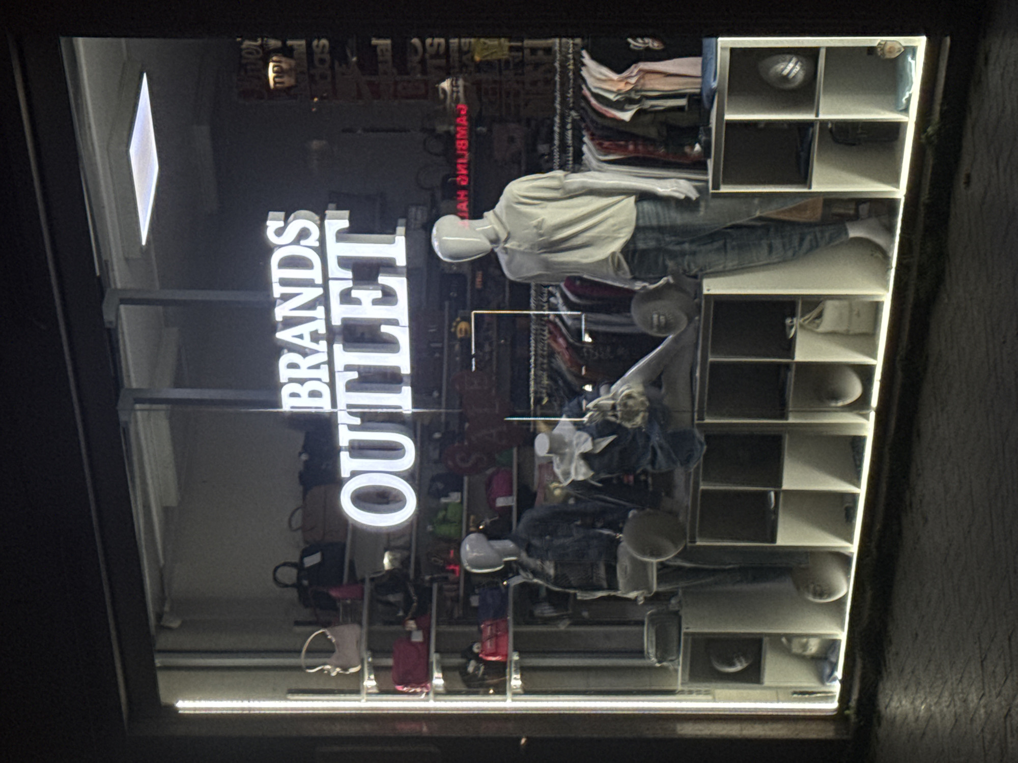 A brightly lit storefront window in what is likely Varna, Bulgaria, displays the illuminated words "BRANDS OUTLET" in stacked formation. Two mannequins, dressed in casual attire, are situated amidst clothing racks and stacked cube shelves containing various objects, including what appear to be mannequin heads. The store’s window reflects the surroundings and adds to the overall visual complexity of the scene.