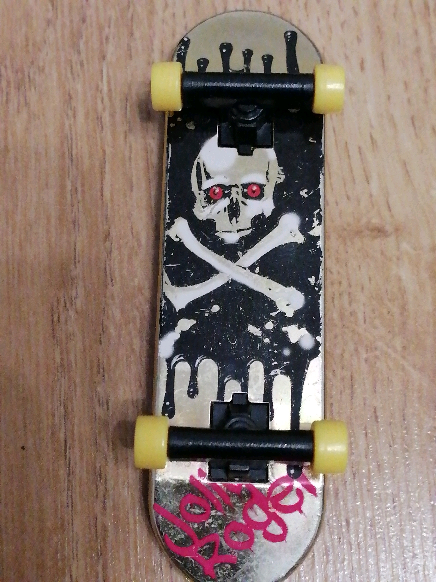 The image displays a close-up, overhead view of a fingerboard, a miniature skateboard. The fingerboard has a metallic gold deck with black trucks and yellow wheels. The underside of the deck features a graphic of a skull and crossbones with red eyes, set against a black background with white splatter effects and dripping black paint. Pink graffiti spelling out "Lil Roger" is visible on the tail end of the deck. The fingerboard rests on a wooden surface, likely a table or floor, with a visible wood grain pattern. The lighting appears to be from above, casting faint shadows. There is no indication of the location being in Kaliningrad, Russia, or any other specific city. The image is a still shot with no discernible action or interaction.
