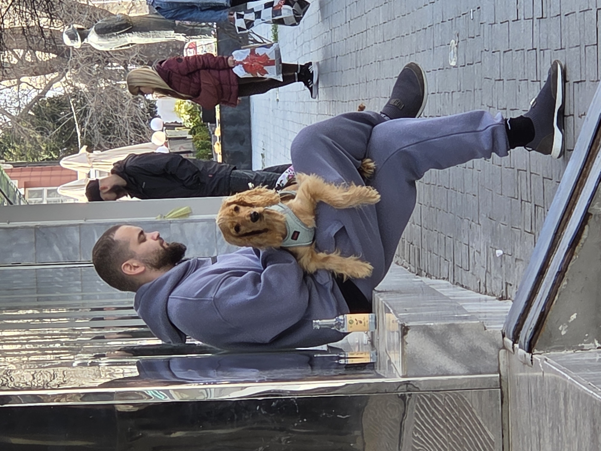The image shows a man sitting on a low, metallic structure on a paved street in Varna, Bulgaria. He wears a dark blue hooded sweatshirt and light blue sweatpants, along with black socks and dark tennis shoes. A golden-colored Cocker Spaniel dog wearing a blue harness rests on his lap. The man appears to be interacting affectionately with the dog. In the background, other people are visible, including a woman in a maroon jacket carrying a checkered shopping bag, and a man in a black jacket and hat. Trees without leaves are also in the background. The low, metallic structure has a clear liquid bottle placed near the man's seat. The location appears to be an outdoor public space within Varna, Bulgaria.