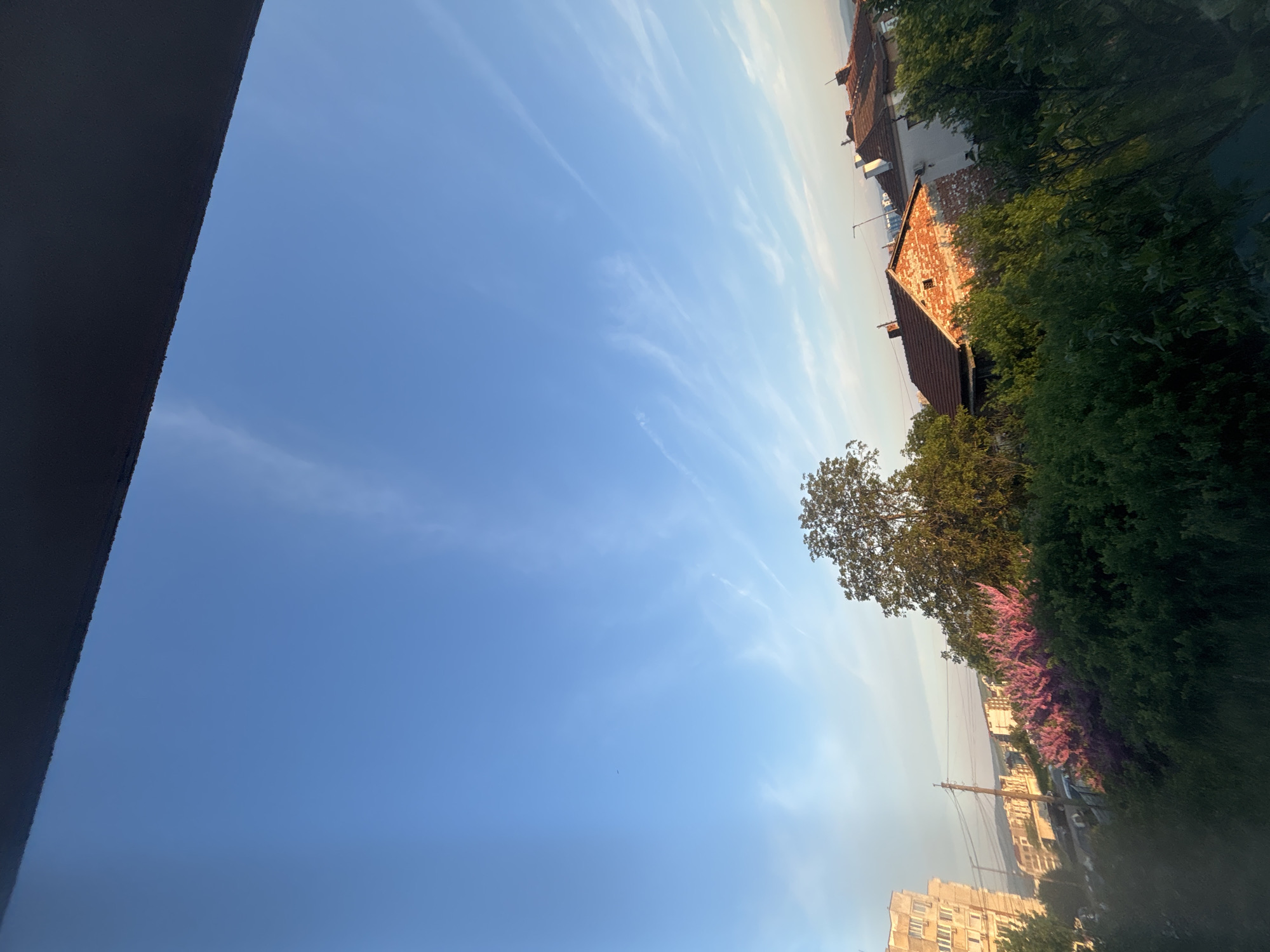 The image presents a view from an elevated position, possibly a high-rise building, in what appears to be a residential area of Burgas, Bulgaria. The foreground is dominated by a vibrant blue sky streaked with wispy cirrus clouds. A portion of the building from which the image is taken is visible in the upper-left corner.
In the midground, a row of buildings with red-tiled roofs and brick facades are partially obscured by lush green foliage. A scattering of trees and flowering bushes add a natural element to the urban landscape.
In the background, a coastline stretches out towards the horizon, possibly indicating the proximity to the Black Sea. Utility poles and buildings further suggest an urban setting.