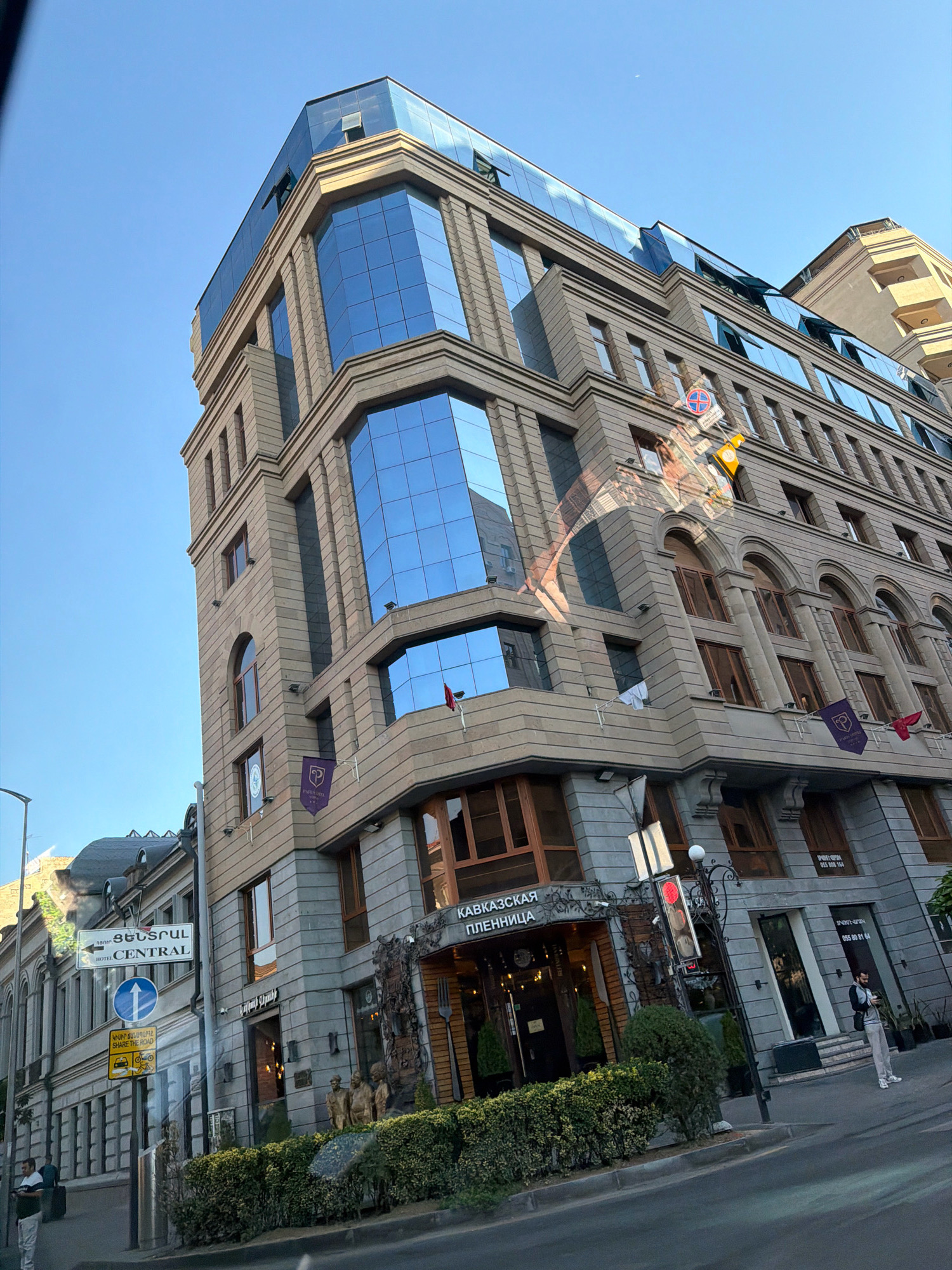 This is a low-angle, eye-level, full shot of a modern building with a glass facade on a city street in Yerevan, Armenia. The building has multiple stories, with the upper floors featuring large, blue-tinted windows. The lower floors are made of stone and have arched windows. There is a sign above an entrance that reads "КАВКАЗСКАЯ ПЛЕННИЦА" (Kavkazskaya Plennitsa) in Russian, which translates to "Caucasian Captive." Below this sign, there is another sign with a phone number. To the left of this entrance, there is a street sign with Armenian text pointing to "CENTRAL HOTEL". Traffic lights and various other street signs are visible. Several people are present on the sidewalk. One person in a white shirt and pants is standing near the entrance, holding a phone. Another person is walking towards the left of the frame. A third person is on the sidewalk near the far left. The sky is clear and blue. The image captures a section of the street with the prominent building as the main subject.