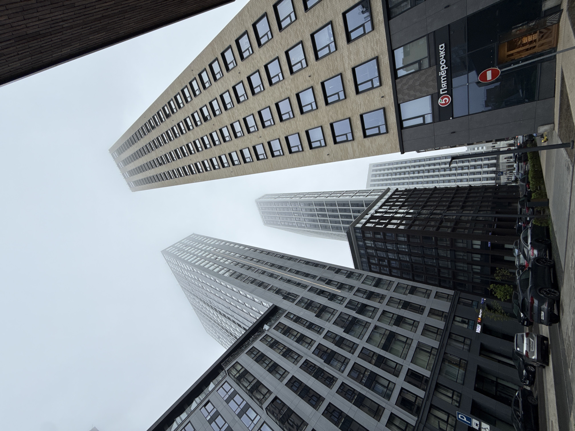 The image captures a low-angle view of several high-rise buildings in Moscow, Russia, obscured by a heavy fog or mist. The buildings exhibit modern architectural styles with numerous windows, and they appear to be constructed from a mix of materials, including glass and concrete. One building prominently features a "Пятёрочка" (Pyaterochka) supermarket logo. Several parked cars are visible along the side of the road, and the overall atmosphere is muted due to the foggy conditions.