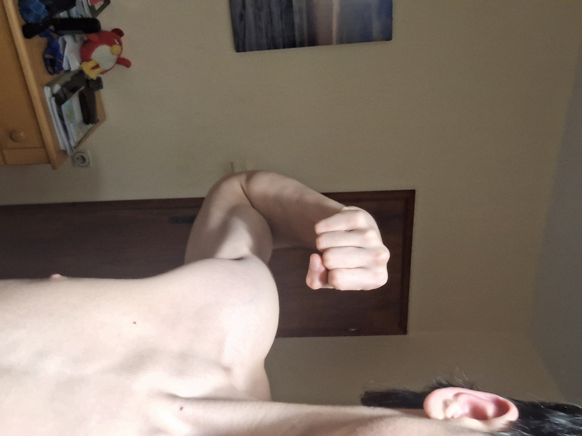 A low-angle shot focuses on a person's physique, specifically the flexed left bicep and clenched fist. The person is fair-skinned with dark hair visible near the ear. Background elements include a dark wooden door, a beige wall with a rectangular artwork, and a cabinet with items such as a red plush toy and papers. The location of the photograph is not discernible from the image.