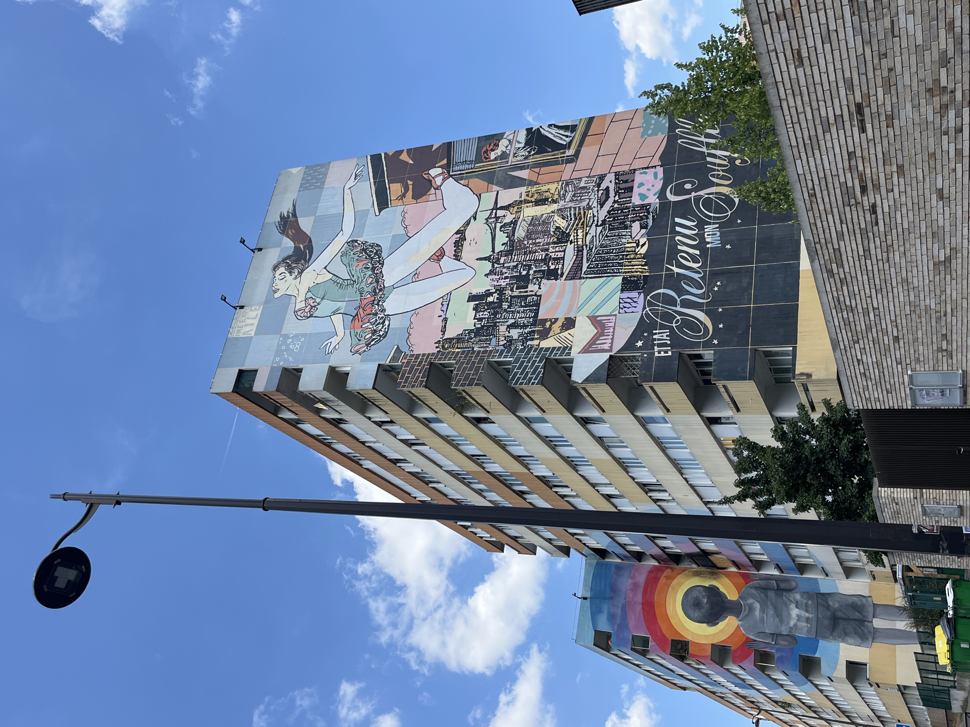 The image depicts a sunny day in an urban environment with a clear blue sky and scattered clouds. In the foreground, a dark gray lamppost stands tall, angled slightly to the right. Behind it, two multi-story apartment buildings are visible. The building on the right is adorned with a large mural featuring a stylized depiction of a woman in a ballet outfit, appearing to leap or dance, with a cityscape below her. Text in cursive script reads "ET J'AI RETENU MON SOUIFFLE". The building on the left displays a vibrant mural of a young boy standing in front of a rainbow, with the sun or a glowing orb behind him. The buildings have multiple floors with visible windows and balconies. A brick wall runs across the bottom of the image, partially obscuring green garbage bins. The scene is likely in Paris, France, given the architectural style of the buildings and the prevalence of street art in certain Parisian neighborhoods.