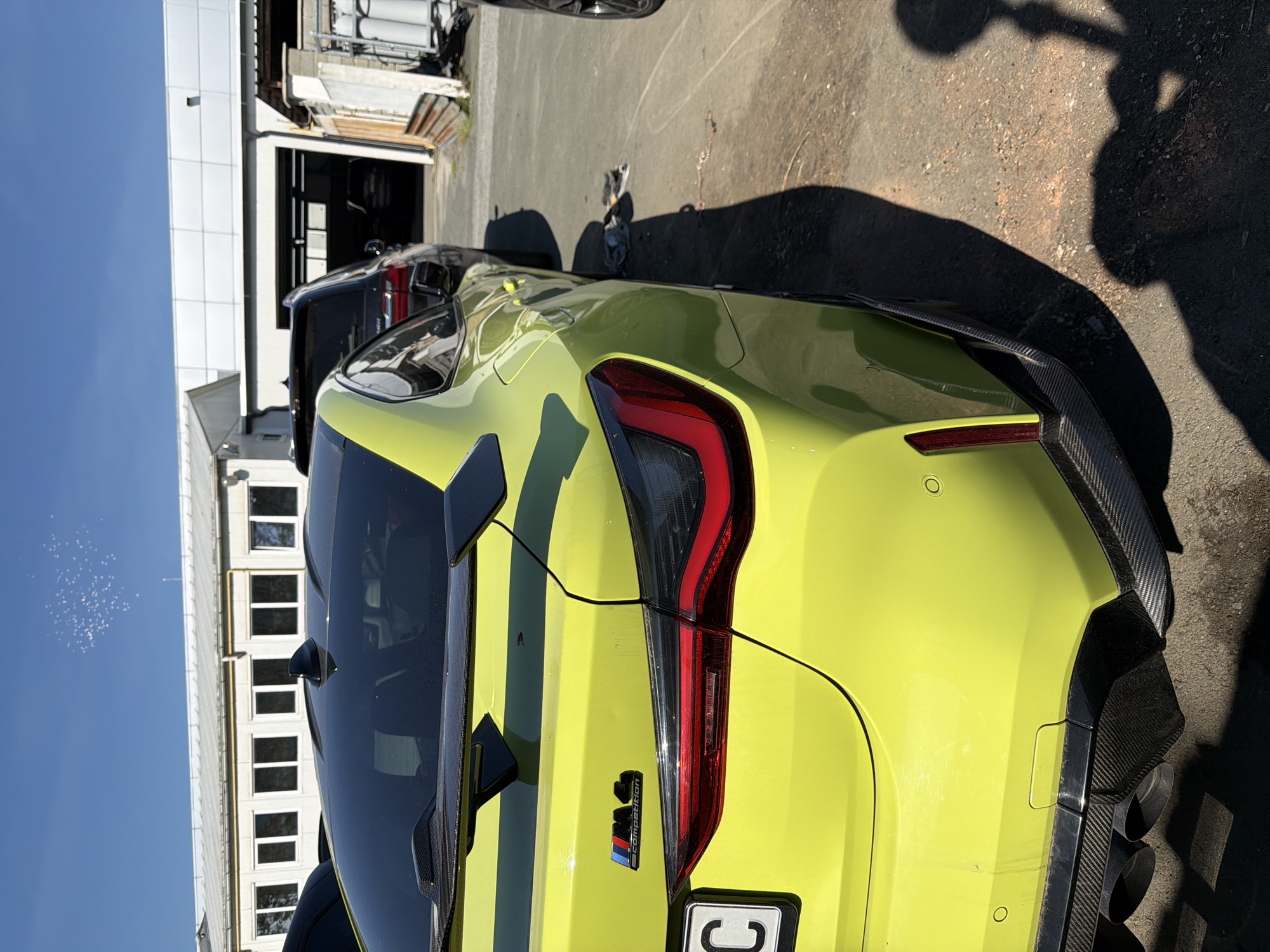 The image shows the rear of a yellow BMW M4 Competition parked outdoors on an asphalt surface. The car has black racing stripes and carbon fiber accents. A black SUV is parked behind it. In the background, there is a white building with windows and a garage-like structure. The presence of a European license plate suggests a location in Europe, and while Burgas, Bulgaria, cannot be definitively determined, it is a plausible location given the style of building and car. There is a shadow of a person or figure visible on the asphalt.