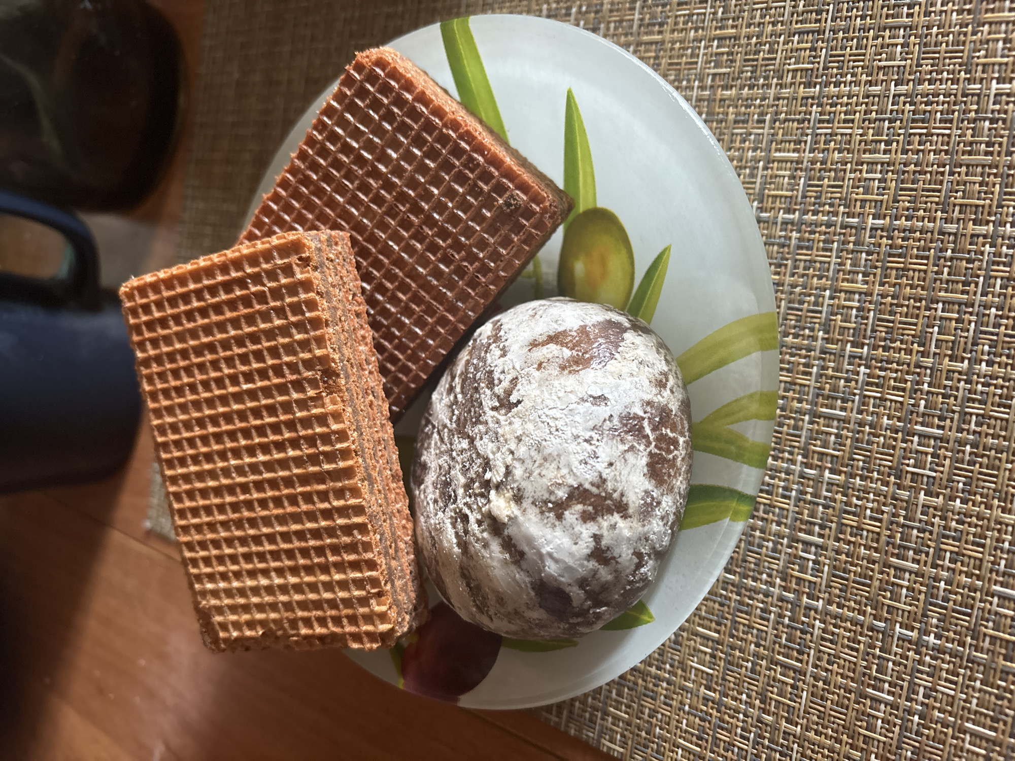 The image shows a plate with two square waffle wafers and a round gingerbread cookie. The plate has a white base with painted green olive branches and olives around the edges. The waffles are light brown and chocolate brown, with a visible grid pattern. The gingerbread cookie is coated in a white, sugary icing. The plate rests on a woven mat with a mix of light and dark brown tones. The location of Chaikovsky, Russia is not discernible from the image content.