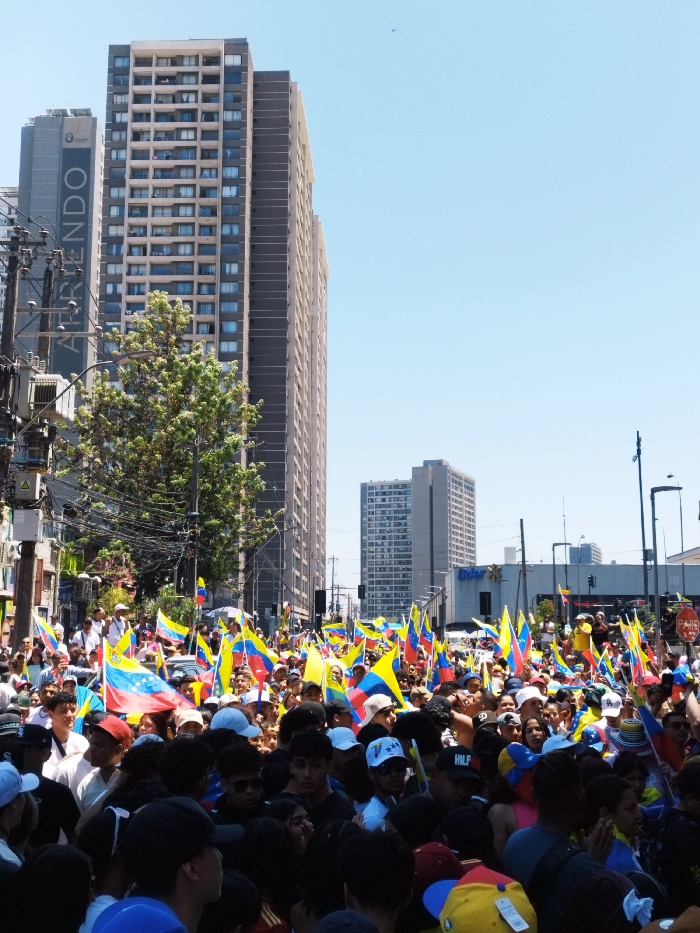 The image depicts a large daytime gathering in a dense urban environment within Santiago, Chile. Hundreds of people are tightly packed in the foreground and mid-ground, extending across the frame, indicating a public assembly or demonstration. Many individuals in the crowd are holding Venezuelan flags, characterized by horizontal yellow, blue, and red stripes with an arc of eight white stars on the blue band. Participants are predominantly adults and young adults, dressed in casual attire suitable for warm, sunny weather.
In the background, a street or wide avenue is flanked by multiple high-rise buildings. On the left, a tall, multi-story building prominently displays "ARRIENDO" (For Rent) written vertically along its side. Further down the street on the right, another commercial building is visible with a partially obscured "Lider" logo, identifying it as a Lider supermarket, a chain common in Chile. Overhead utility poles and complex networks of power lines are visible on the far left. A large green tree stands in the mid-ground, partially obscuring the buildings behind it. The sky is clear and light blue, indicating bright, sunny weather. A "PARE" (STOP) sign is partially visible on the right.