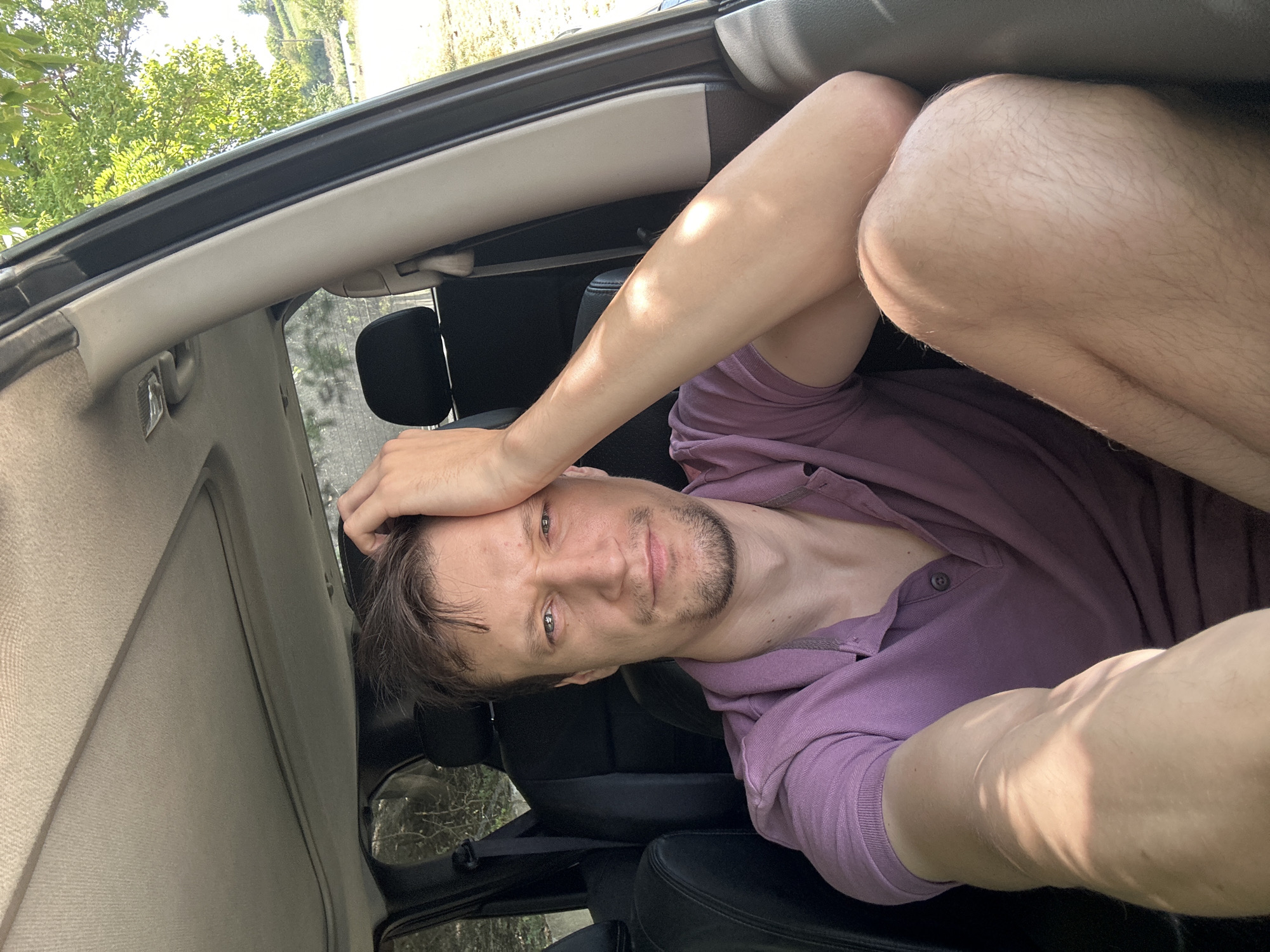 A man with short brown hair, a light beard, and a purple polo shirt is sitting in the driver's seat of a car. He is looking at the camera and has his right hand on his forehead. Sunlight is shining on his arm and leg. The interior of the car is visible, including black leather seats and a sunroof. Outside the car, trees and a distant hillside are visible, suggesting a rural or semi-rural location. The scene appears to be set in Makiv, Ukraine.