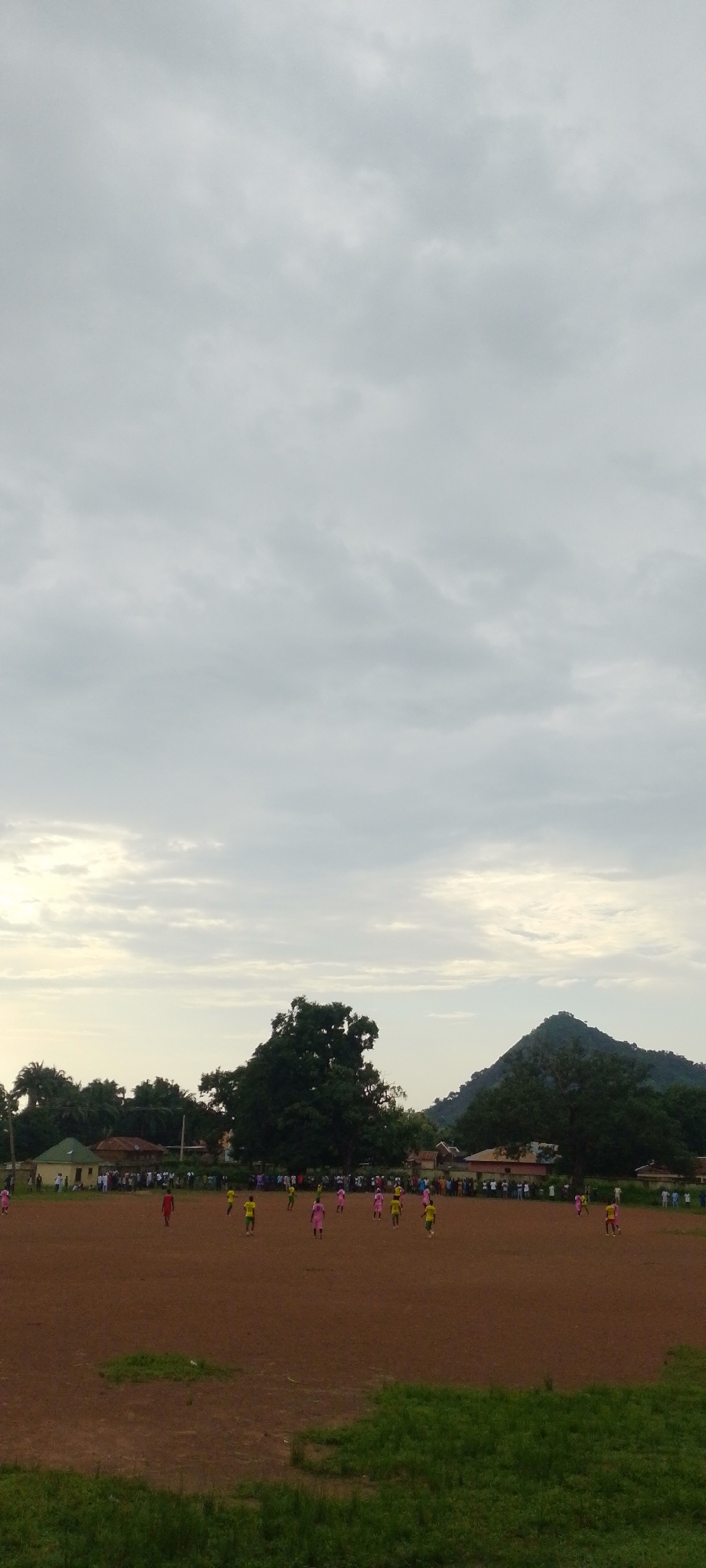 This media file shows an outdoor scene with a football match in progress. The setting is a dirt football field, with a large crowd of spectators gathered along the sidelines. In the background, there are buildings and lush green trees, with a prominent hill or mountain in the distance. The sky is overcast with clouds, suggesting it might be late afternoon or early evening, or perhaps a day with unsettled weather.
The primary subjects are the people playing football on the field. There appear to be two teams, distinguishable by their colored jerseys. Players in pink and yellow/green jerseys are visible, actively engaged in the game. The spectators are also a significant subject, forming a dense line along the edge of the field, indicating their interest and support for the match. The overall atmosphere seems to be one of communal gathering and engagement in a sporting event.
Notable details include the worn appearance of the dirt field, common in many informal or community sports grounds. The presence of varied vegetation and the distant geographical feature suggest a rural or semi-rural location. The cloudy sky contributes to a somewhat subdued lighting, without strong shadows. No text is visible in the image. The event is clearly a football match, likely a community or local league game, given the size of the crowd and the informal nature of the field.