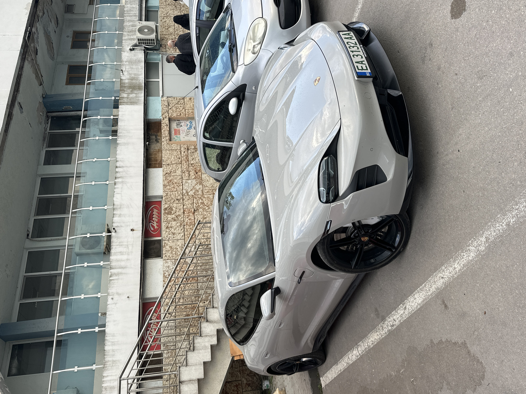 The image shows two cars parked in front of a building with a glass-enclosed staircase. The closer car is a light gray Porsche Taycan with a Bulgarian license plate "EA 3132 AA." Behind the Porsche is a silver hatchback. Two people stand near the hatchback. The building is multi-storied, with a weathered facade. A "Happy" restaurant sign is visible on the building's facade. The location is Varna, Bulgaria.