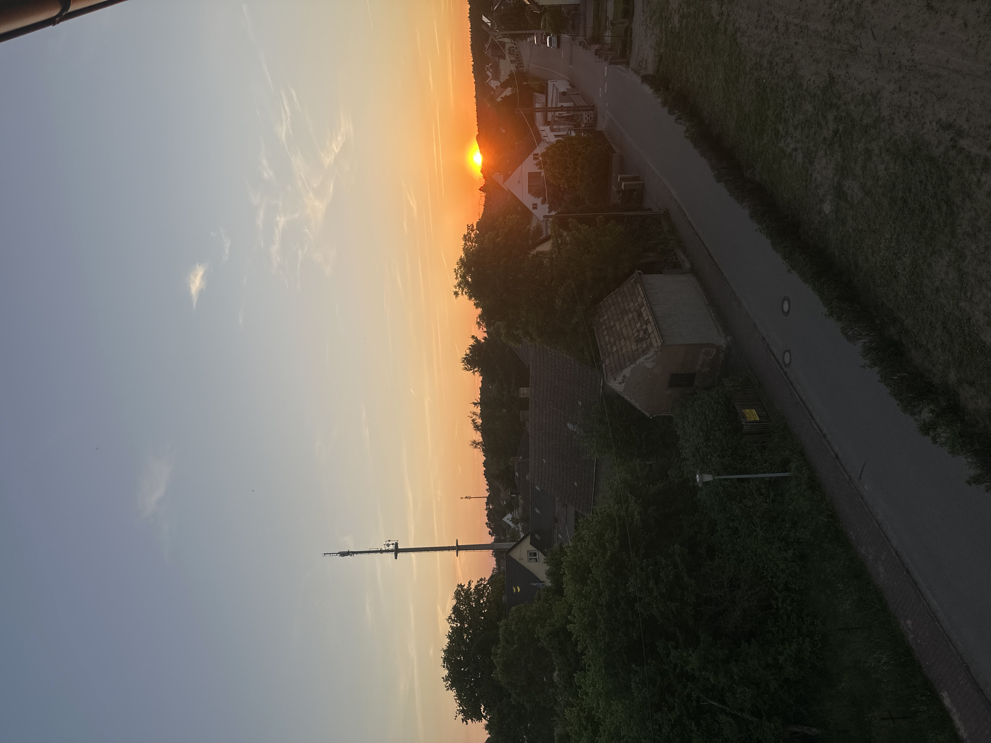 The image depicts a sunset over Arzberg, Germany. The sun is setting on the horizon, casting an orange glow over the sky and illuminating the surrounding landscape. Below the horizon, there are buildings of what look like a village, fields, trees, and a road. A tall communications tower stands in the distance, near the center of the picture.