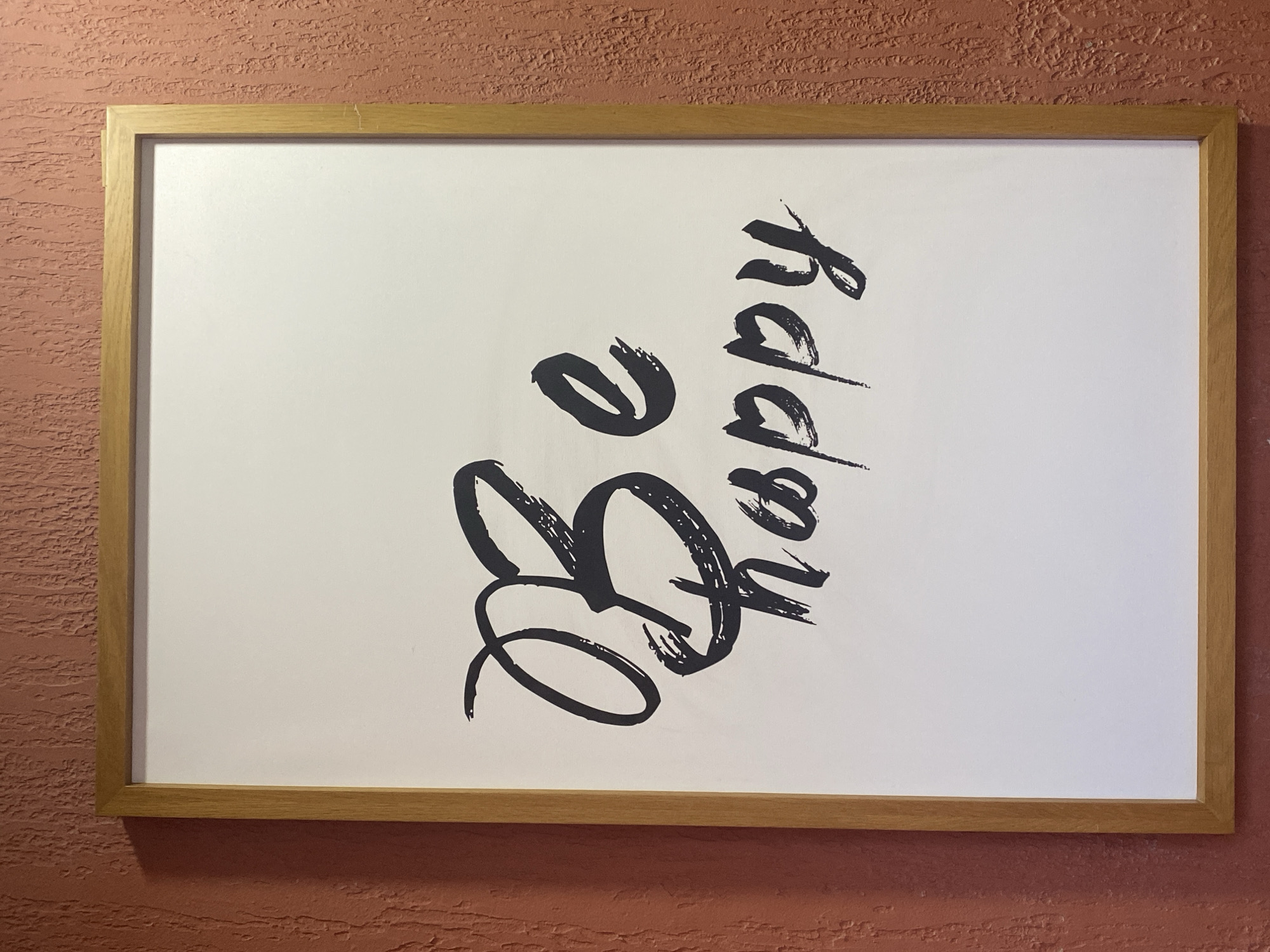 A framed piece of art with the words "Be happy" written in a calligraphic font is displayed on a textured, reddish-brown wall. The frame is made of light-colored wood. The artwork itself is white with black lettering. The overall scene is static and appears to be an interior decoration. There are no people or actions depicted.