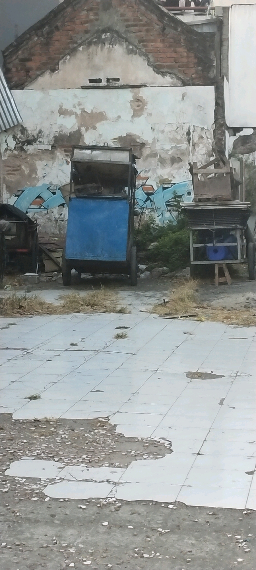The image depicts an outdoor scene with two carts in front of a dilapidated building. The building has peeling paint and graffiti on its walls, with exposed brickwork visible. The ground is paved with cracked tiles, and some areas are covered with debris and dry grass.
The cart on the left is a metal frame with a blue panel at the front, and a metal roof. The cart on the right is also a metal frame, with a wooden structure on top and a glass-enclosed area at the bottom, containing a blue bucket and a wooden stool. There are no people visible in the image. The overall impression is one of abandonment and decay. This scene is located in the city of Surabaya, Indonesia, as evidenced by the architecture and the style of the carts, which are commonly used by street vendors in the region.