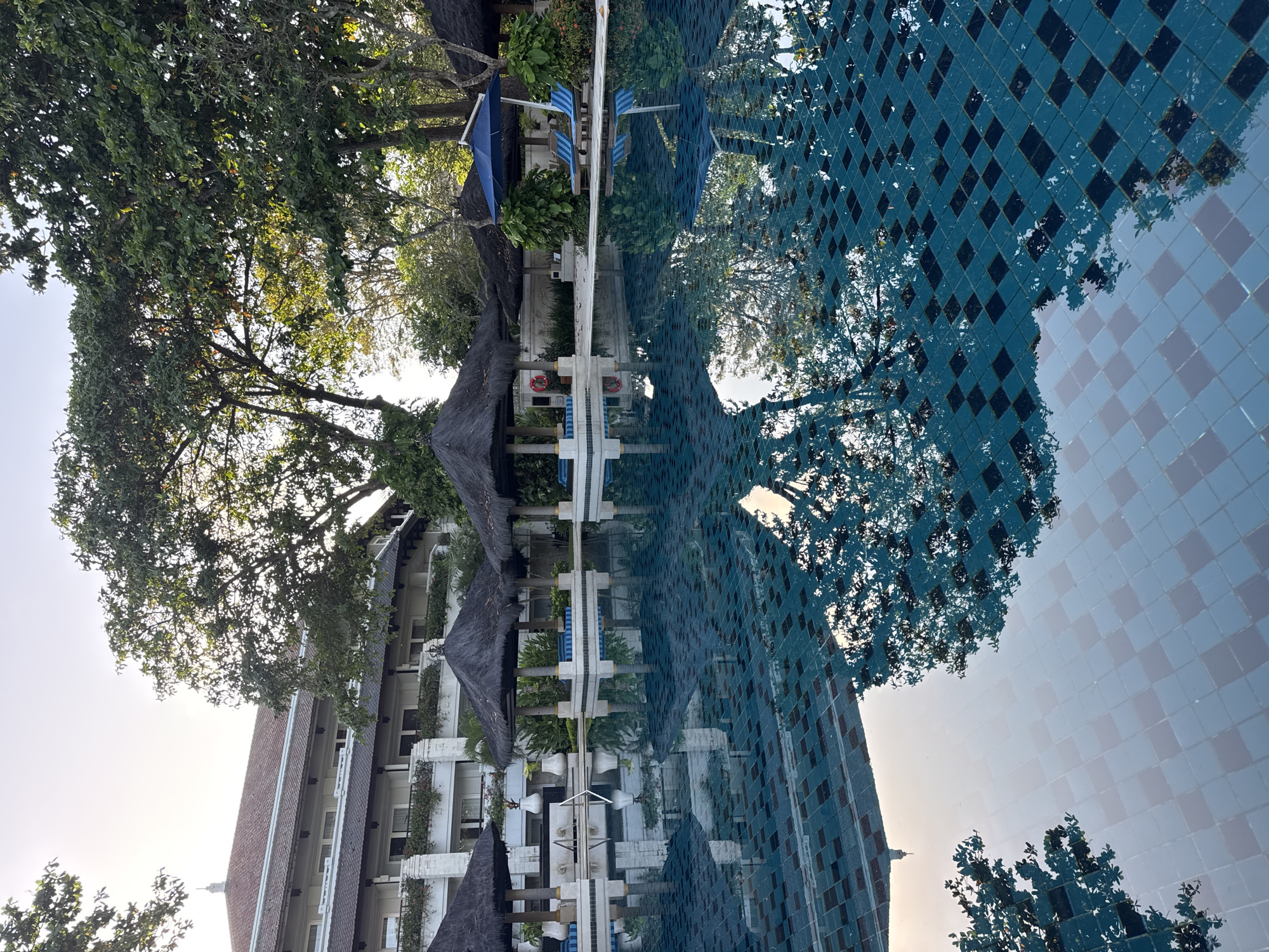 This image depicts a view of a resort or hotel in Jimbaran, Indonesia, possibly reflecting in a swimming pool. The building features multiple stories with white walls, balconies, and a reddish-brown roof. Some thatched-roof structures are present, likely providing shade near the pool. The presence of blue umbrellas and lounge chairs implies a relaxation area. The reflection in the water shows a pattern of blue and black tiles, along with the distorted image of the building and surrounding trees. The lighting indicates it's likely daytime.