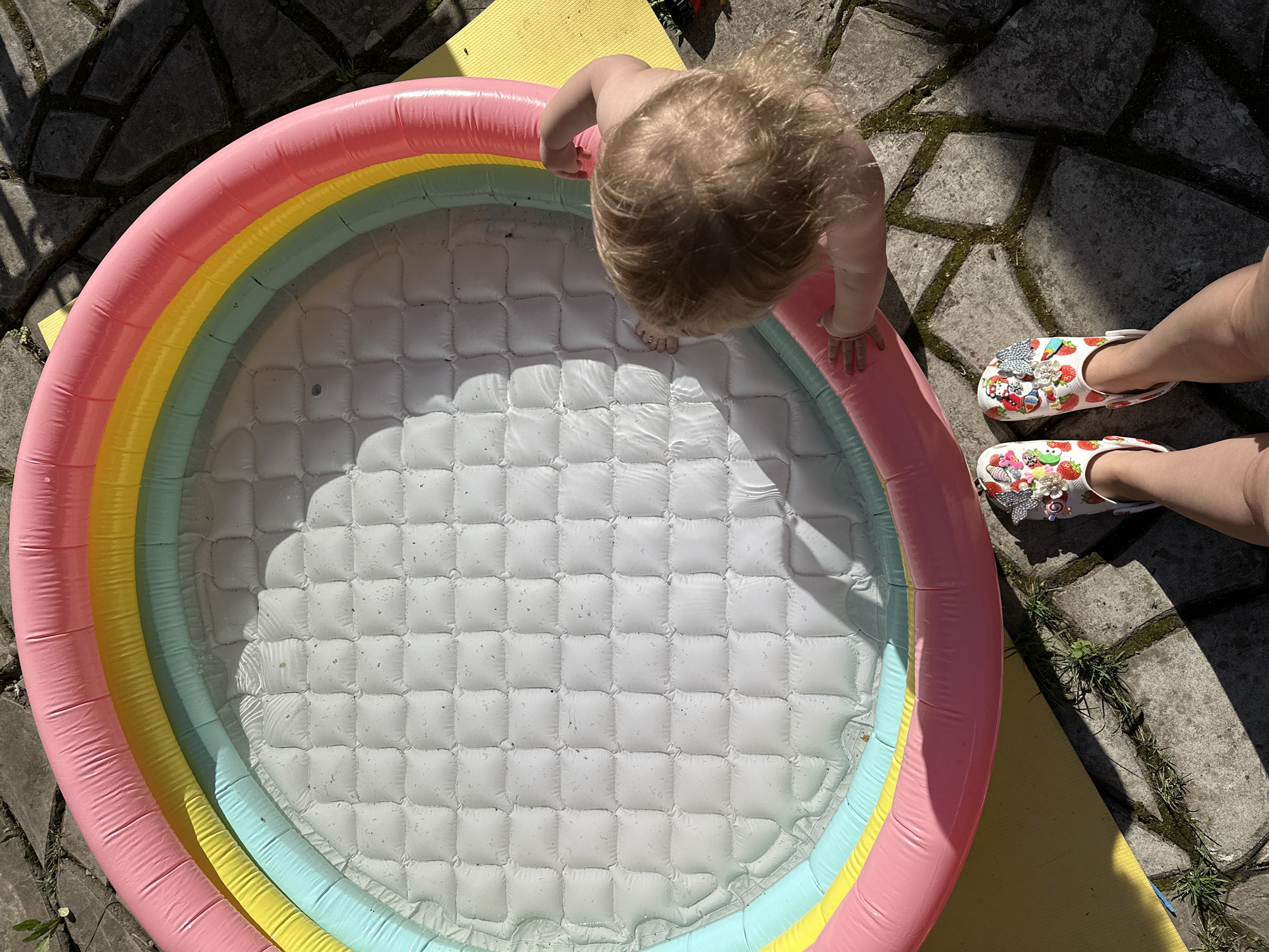 An overhead shot shows a child leaning into a rainbow-colored inflatable pool that is partially filled with water. The pool is situated on a yellow mat. The child appears to be blond. Next to the pool are the legs of a person wearing white clogs decorated with strawberries and decorative charms. The pool sits on a gray stone surface overgrown with weeds in some spots. There is no evidence to suggest the image was taken in Nizhny Novgorod.