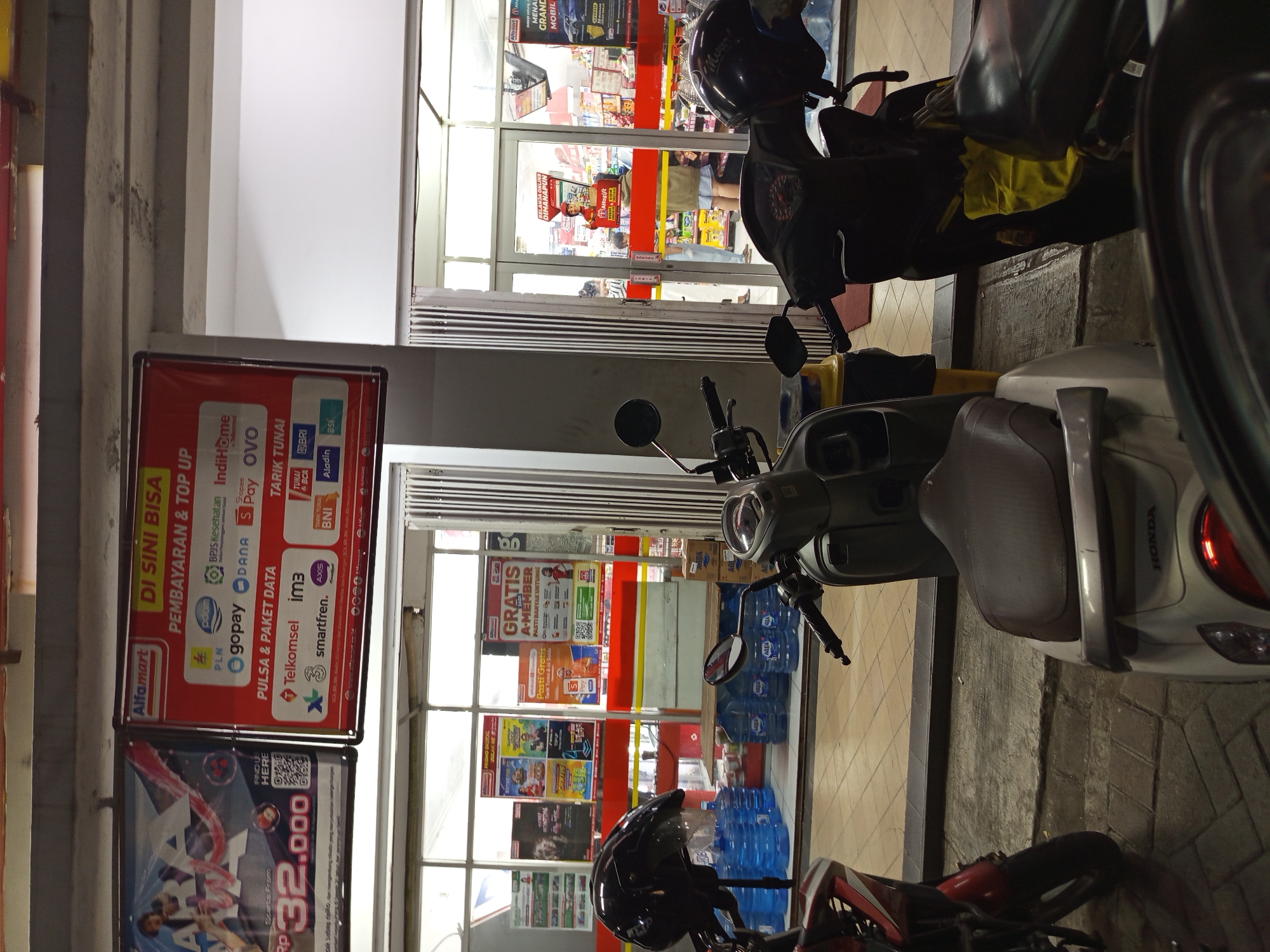 This is a photograph of the exterior of an Alfamart convenience store, likely located in Gemurung, Indonesia, as indicated by the signage. The store is viewed from a low angle, with several motorcycles parked in front of its entrance. The entrance consists of a glass door with vertical metal bars and a glass window, revealing the interior of the store, which is stocked with various goods. Inside, a person in a yellow shirt and blue shorts is standing near the checkout counter, interacting with another person. Above the entrance, a large red and white banner displays promotional information in Indonesian, including "DI SINI BISA PEMBAYARAN & TOP UP" which translates to "HERE YOU CAN PAYMENT & TOP UP." This banner lists various payment methods and services available, such as PLN, Poom, BPJS Kesehatan, IndiHome, GoPay, DANA, OVO, Telkomsel, IM3, Smartfren, AXIS, and different bank logos like BRI and BNI. To the left of the main entrance, a smaller banner is partially visible, featuring a woman and text indicating a price of "Rp 32.000". Several large blue water dispensers are stacked in front of the store's windows, along with boxes of Aqua water bottles. Two helmets, one black and one white, are placed on the handlebars of the parked motorcycles. The ground in front of the store is paved with tiles and concrete. The overall scene depicts a typical retail environment with motorcycles parked outside, suggesting a common mode of transportation for customers in the area.