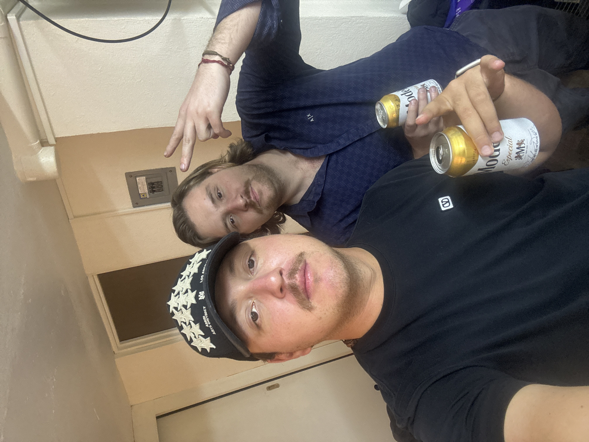 Two men are posing for a photo indoors. The man in the foreground is wearing a black t-shirt and a black cap with stars and text that reads "ARTS DISTRICT AMIRI 101 LOS AN AVEZA". He has a mustache and a slight stubble. The man in the background is wearing a patterned navy blue shirt. They are holding Modelo Especial beer cans. The background includes a wall, a window, and an electrical panel. The location is inside a building, possibly in Huixquilucan de Degollado, Mexico.