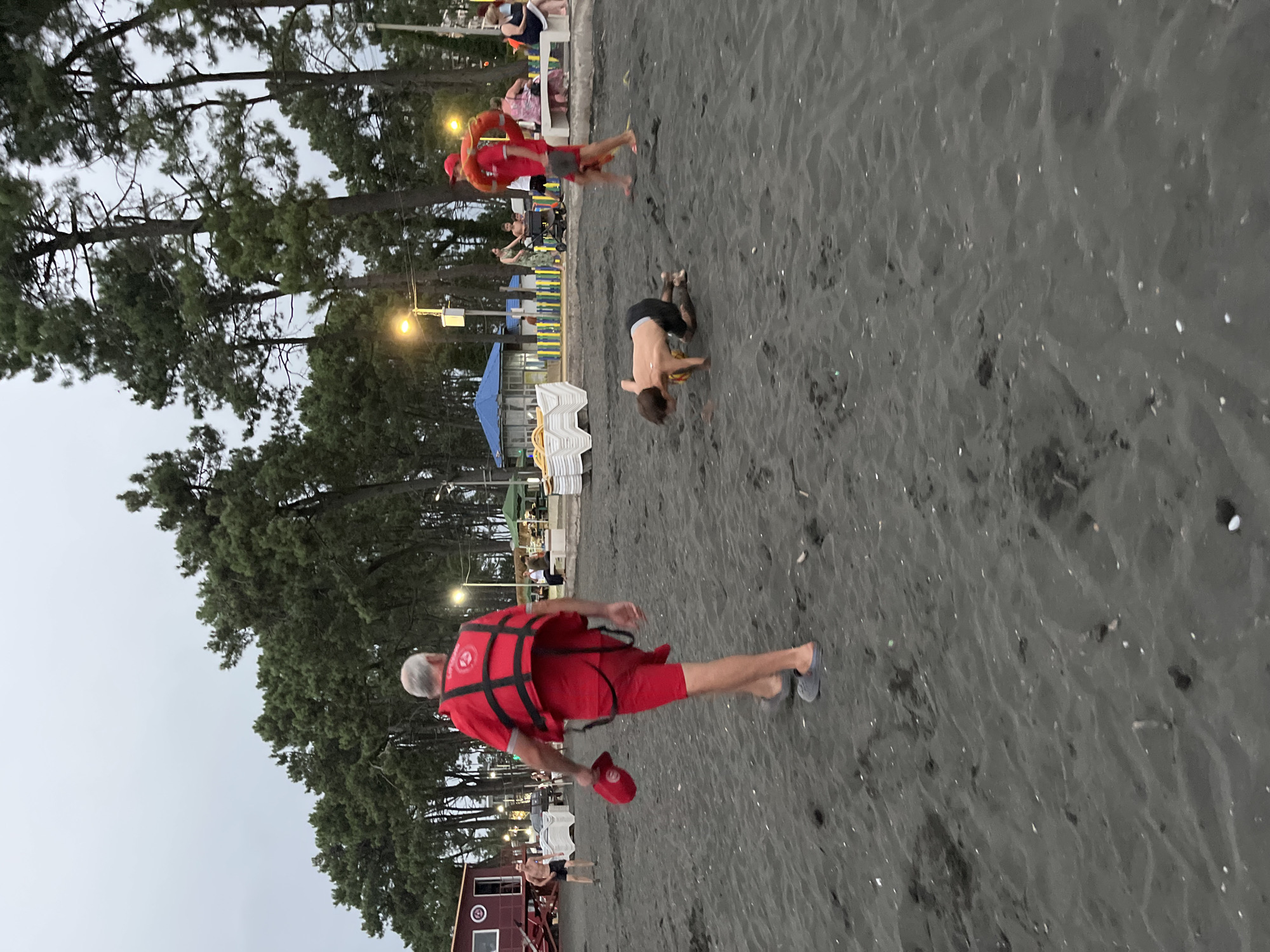 This is a photograph taken on a beach in Ureki, Georgia. The foreground is dominated by dark sand, with a few scattered white shells and small debris. In the mid-ground, a man wearing a red vest and shorts, with a red cap in his hand, walks away from the camera. He is also wearing grey sandals. To his left, a young boy is on his hands and knees on the sand, looking down. Further in the mid-ground, another person in a red vest and shorts, holding a red ring, walks from left to right. In the background, there are several pine trees and some buildings, including a blue and white structure. There are also people sitting on a bench and a child in a stroller. The sky is overcast, and there are streetlights visible. The overall impression is of a beach area with a lifeguard presence.