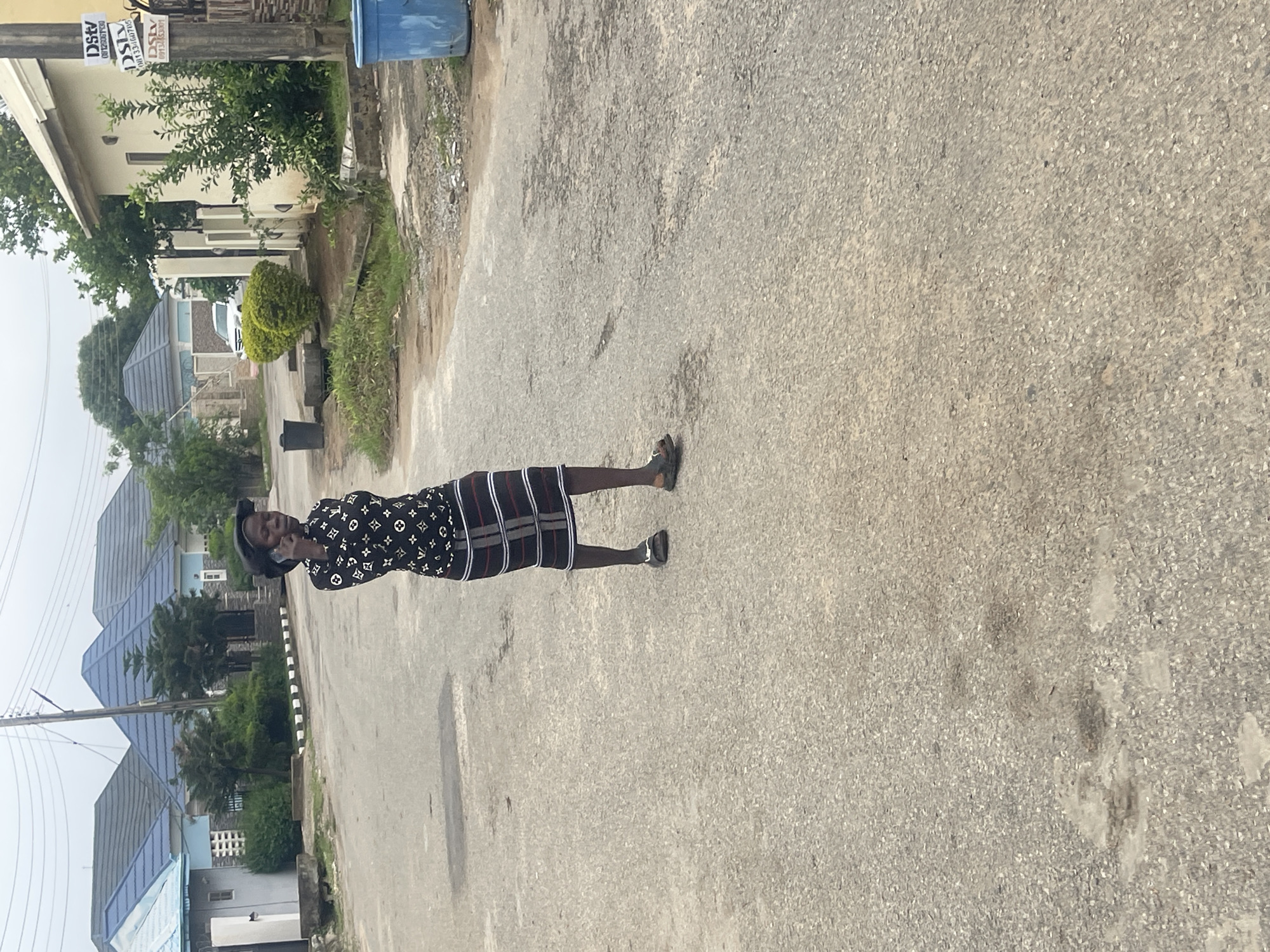 A person stands in the middle of a paved road in Abuja, Nigeria, holding a cell phone to their ear. The person is wearing a black and white patterned long-sleeved shirt and a skirt with horizontal stripes in black, white, and red. They are also wearing dark sandals. The road is lined with houses and lush green trees. In the background, a white SUV is parked on the side of the road. A blue trash bin is visible on the right side of the image, and a black trash can is on the left. The sky is overcast.