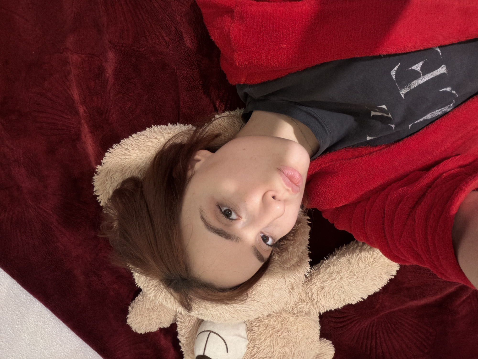 The image displays a young adult female lying on her back, positioned diagonally across the frame, looking directly towards the camera. Her head rests on a light brown plush teddy bear, which appears to function as a pillow or cushion; the bear's head, with a stitched smile and eyes, is visible above her. She has reddish-brown hair, a pale complexion, and her lips are slightly pursed in a pout. She wears a dark gray t-shirt with partially visible light-colored text (appearing to be "LE") and a thin gold-colored necklace. A red plush robe or blanket covers her torso and arms. She is situated on a dark red, textured, plush blanket or bedspread. A white wall or surface is visible in the upper right background. The setting is an indoor, private environment; no specific visual cues within the image identify its precise location within Strasbourg, France.