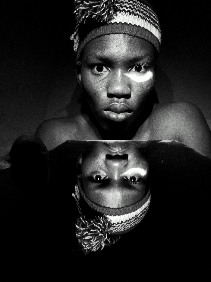 This black and white image depicts a close-up portrait of a single individual, located in Keffi, Nigeria. The subject, appearing to be a young person with dark skin, is positioned centrally in the upper half of the frame, gazing directly forward with wide, attentive eyes and slightly parted lips. A distinct white, roughly oval-shaped marking is visible on the subject's left cheek (viewer's right), extending towards the nose bridge. The individual wears a knit beanie with horizontal light and dark stripes and a prominent pom-pom on top. The upper torso is bare, with shoulders and the upper arms visible. Immediately below the subject, a dark, reflective surface mirrors the individual's head, beanie, and upper torso in an inverted orientation, creating a symmetrical composition. The background is uniformly dark, providing no discernible environmental details. Lighting appears to be directed from above, highlighting facial features and the white cheek mark.