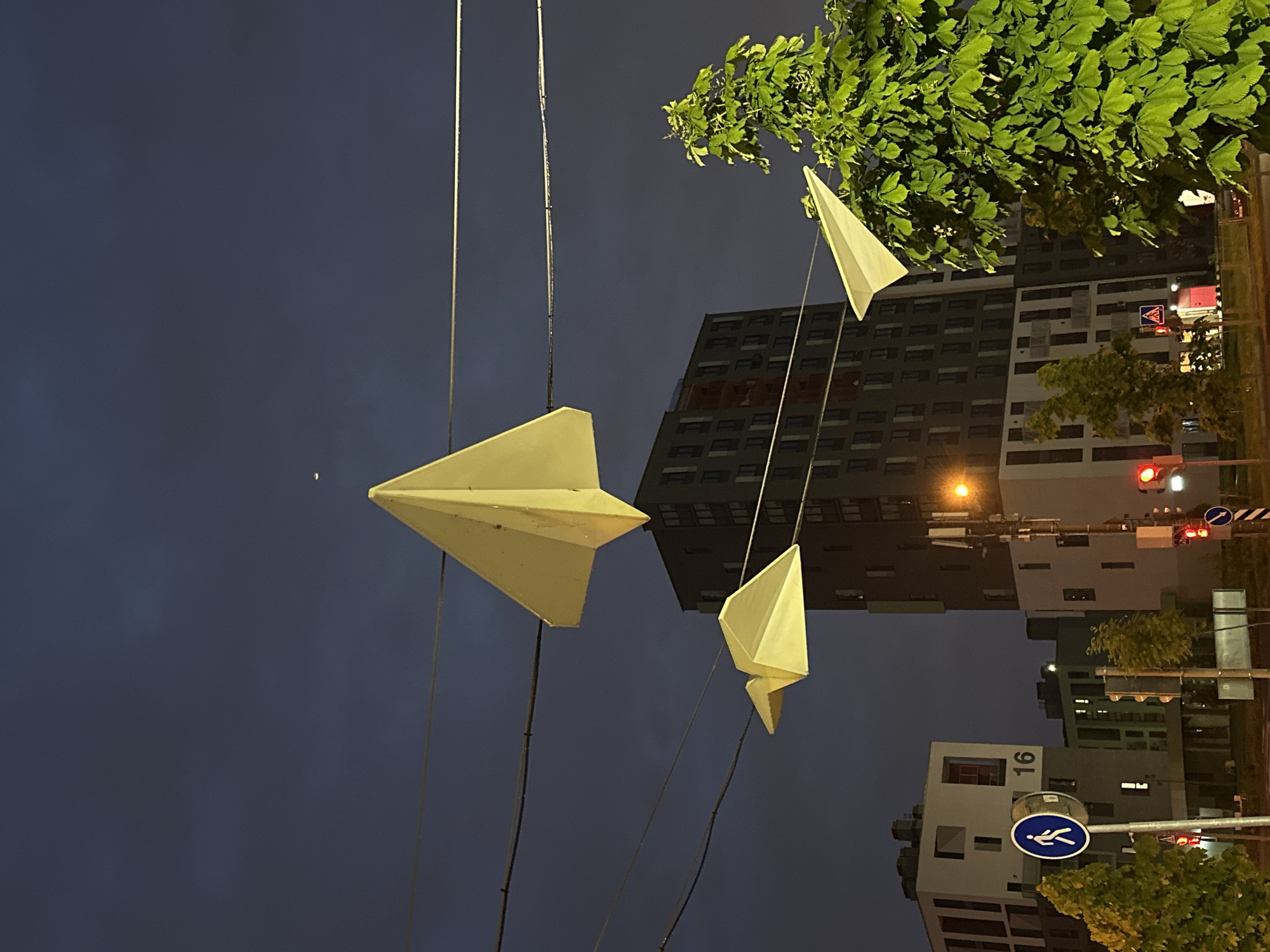 The image depicts an urban scene in the evening or at night, likely in Бараўлянскі сельскі Савет, Belarus. There are three large, white paper airplane sculptures suspended by thin wires, with the night sky as the backdrop. A tall, dark building with many windows is visible behind the sculptures. To the right, a street with traffic lights, pedestrian crossing signs, and other buildings are present. The illumination suggests it is nighttime.