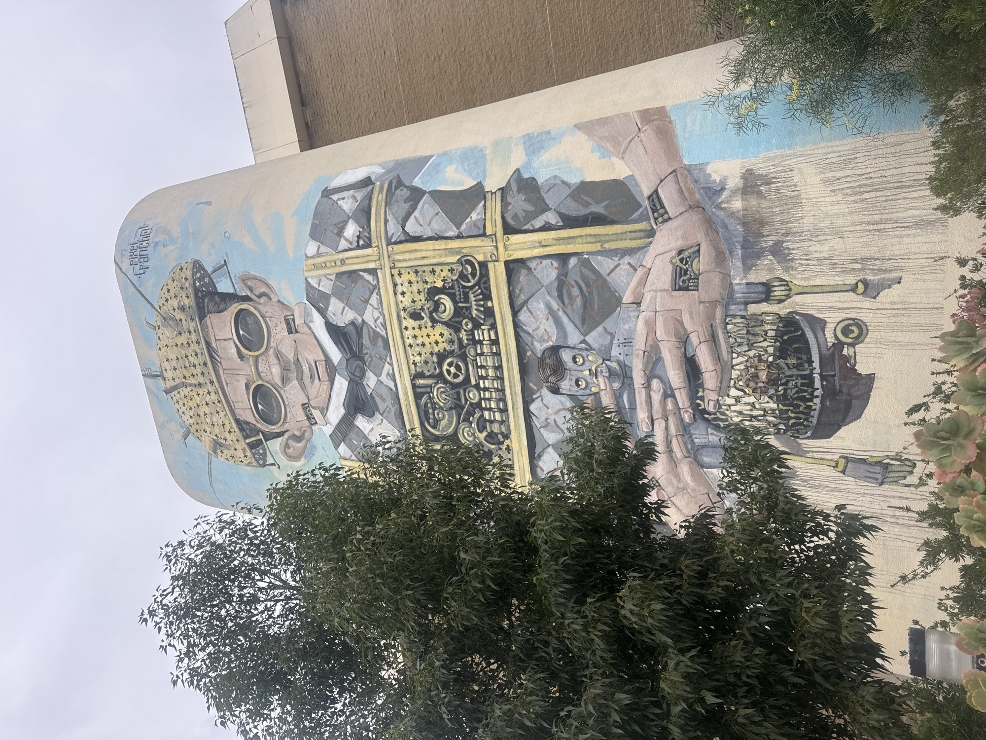 This image shows a large, colorful mural painted on the side of a curved building. The mural depicts a stylized, robotic figure with a patterned shirt, a hat with antennae, and goggles. The figure appears to be holding a smaller robotic head and has mechanical components visible in its abdomen. The building itself is a concrete structure with a curved facade, likely part of a commercial or public building. Vegetation, including a large tree and some succulents, is present in the foreground, partially obscuring the mural. The sky is overcast. The mural is located at the corner of Barrack Street and Wellington Street in Perth, Australia, and is signed "Pixel Pancho."