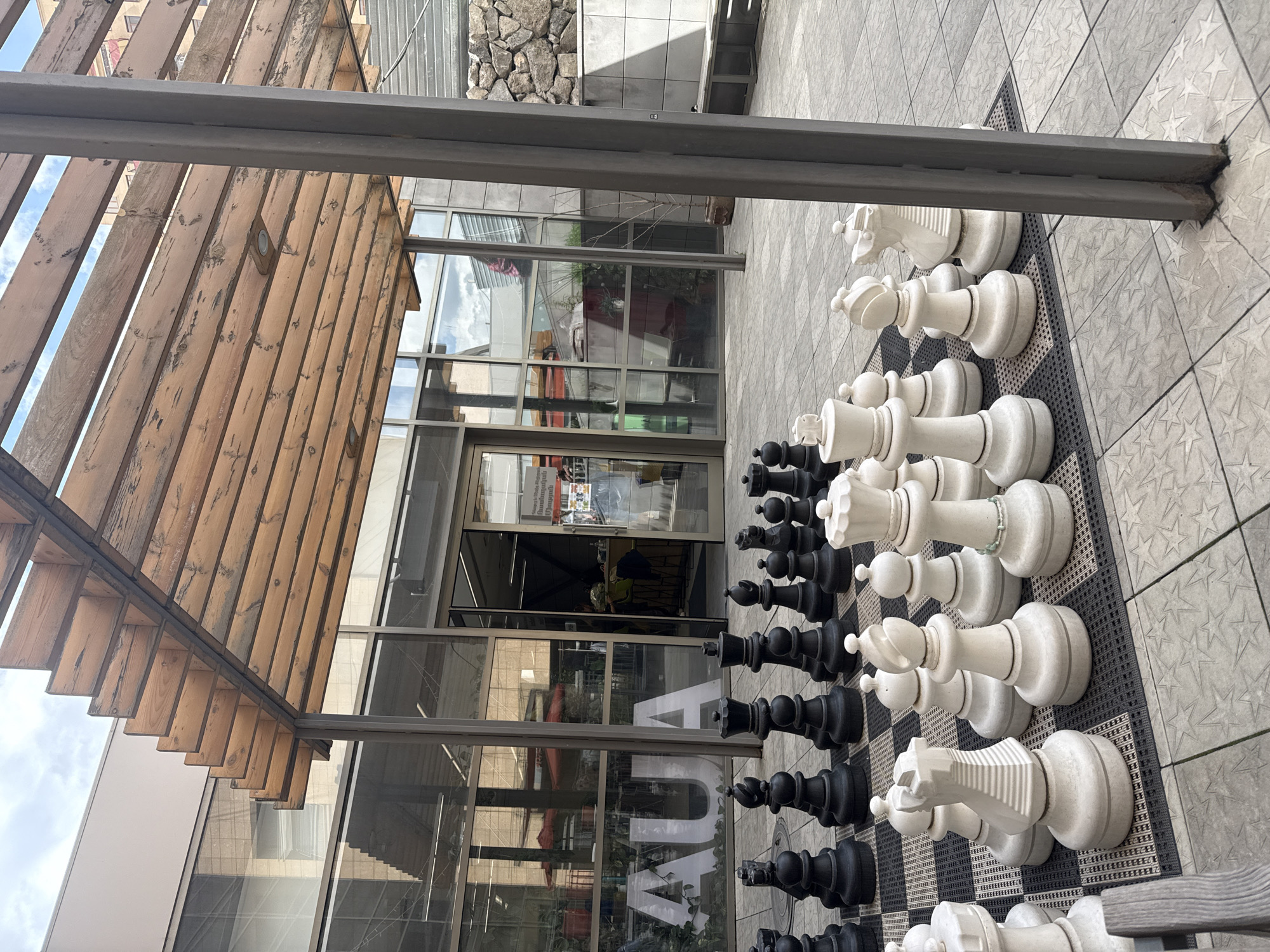 A partially-rotated, eye-level shot depicts a giant outdoor chess set in Yerevan, Armenia. The chess pieces, black and white, are lined up on a checkerboard mat laid over grey square tiles.
Overhead, a wooden pergola structure with a steel frame is visible, partially blocking the view of the building behind the chess set. The building has large glass windows and doors. Through the glass, interior details like plants, furniture, and signage are visible. People are seen inside the building, one with a visible apron. The letters "AUA" are prominently displayed on a window. A stone-clad wall is visible on the building, suggesting mixed architectural styles. The surrounding area is paved with grey tiles, and stairs leading to a different level are seen in the upper right.