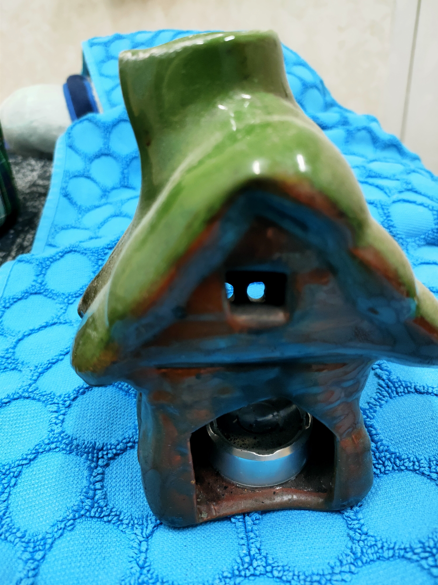 This is a close-up, eye-level photograph of a green ceramic oil or wax warmer shaped like a whimsical house. The object is placed on a textured, bright blue towel. The house has a pointed, green roof and a darker, reddish-brown base that appears to be made of clay or a similar material. There's a small, square opening on the front of the house, revealing two circular holes within. Below this opening, at the base of the house, is a silver-colored metal tea light candle holder, containing a partially burned candle. The lighting in the image suggests it's daytime, and the setting appears to be indoors, possibly a bathroom or a shelf area, given the presence of the towel and what looks like a tiled wall in the blurred background. The overall mood is calm and domestic, with the focus on the decorative object and its potential for creating a pleasant ambiance. No text is visible in the image.
