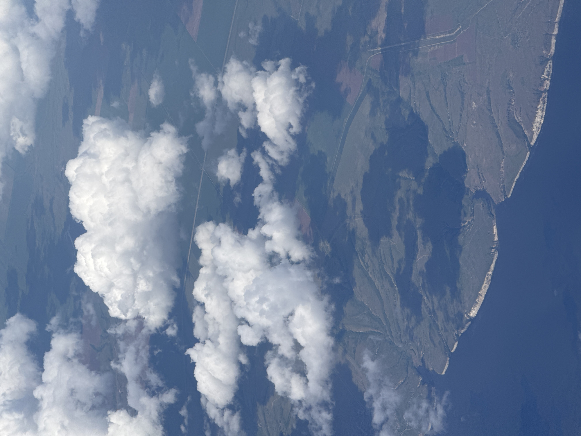The image shows a landscape featuring land, water, and clouds, possibly captured from an aerial perspective. The right side reveals a landmass with a distinct coastline bordering a body of water. The coastline appears to have light-colored cliffs or bluffs. The land itself is vegetated. Clouds are prominently featured on the left side of the image, obscuring portions of the landscape. The presence of fields suggests agricultural activity in the area not covered by clouds. While the image is labeled as Izhevsk, Russia, there is no specific feature visible in the image to confirm the location.