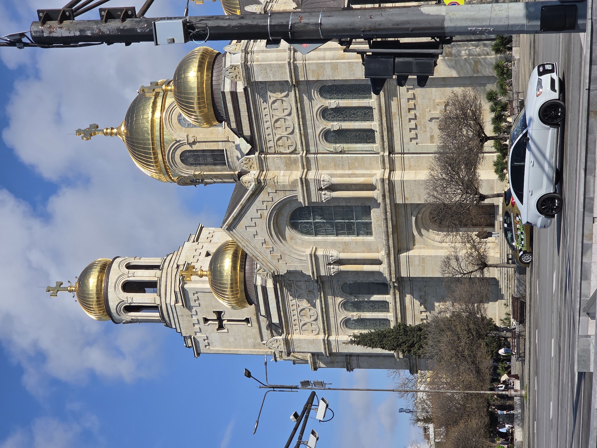 The image shows the Dormition of the Mother of God Cathedral in Varna, Bulgaria, from a street-level perspective. The cathedral, a large stone building with golden domes, dominates the frame. A white car is parked on the street beside it. A few bare trees stand near the cathedral, and groups of people are sitting on benches on the sidewalk. A road runs alongside the building. Streetlights and traffic signals are visible. The sky is blue with some clouds. The image appears to be rotated 90 degrees clockwise.