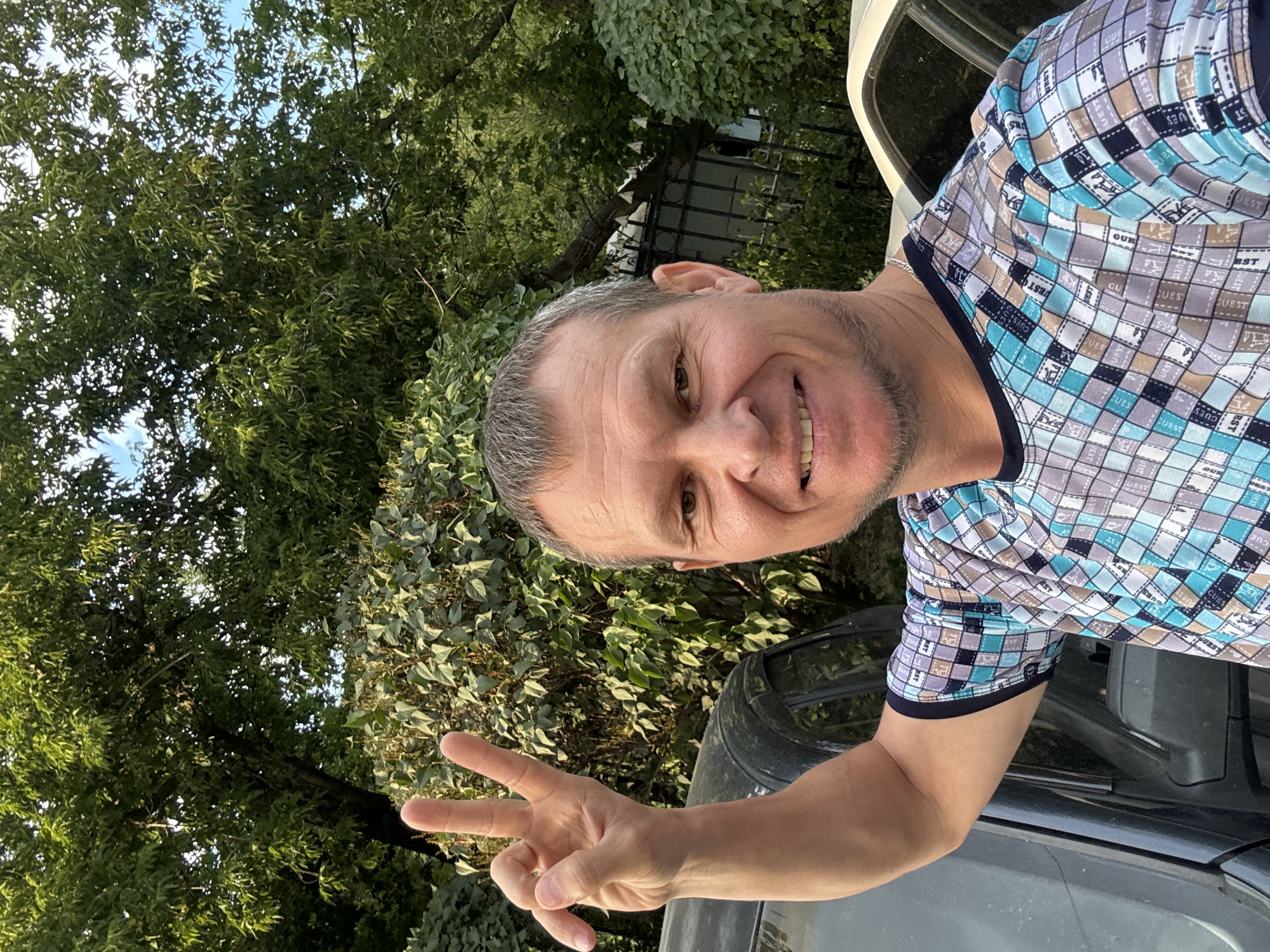 A man with short graying hair and a beard, wearing a t-shirt with a checkered pattern, holds up two fingers in a peace sign. He is standing outdoors, with trees and foliage in the background. He appears to be in front of two cars. The image is a selfie, taken from a low angle. The lighting suggests it is daytime. The specific location in Oskemen, Kazakhstan cannot be determined from the visual information provided.
