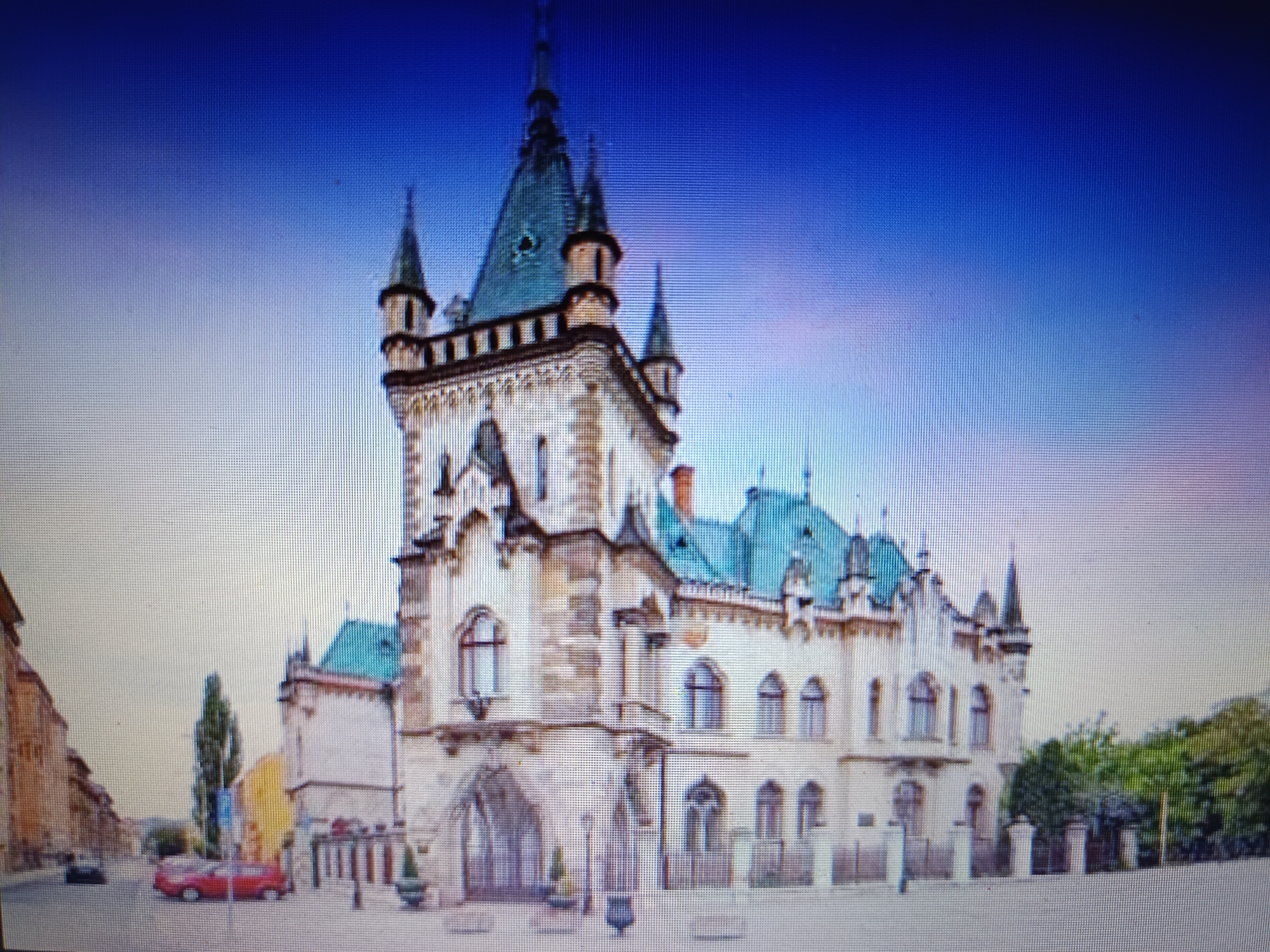 The image depicts the historic Ivanka House (also known as Kasárne/Kulturpark) in Košice, Slovakia, under a bright blue sky. The building features a distinctive neo-Gothic architectural style, characterized by its multiple spires, ornate facades, and arched windows. The primary structure is made of stone and has a light-colored facade. Green-tiled roofs top the building, contrasting with the stone. In the foreground, a street is visible with a red car parked on the right side and a dark-colored car further down the street on the left. Trees and foliage surround the building, indicating a presence of greenery within the urban environment. The image appears to be a photograph, possibly taken from a slight distance, capturing the facade of the Ivanka House and its surroundings.