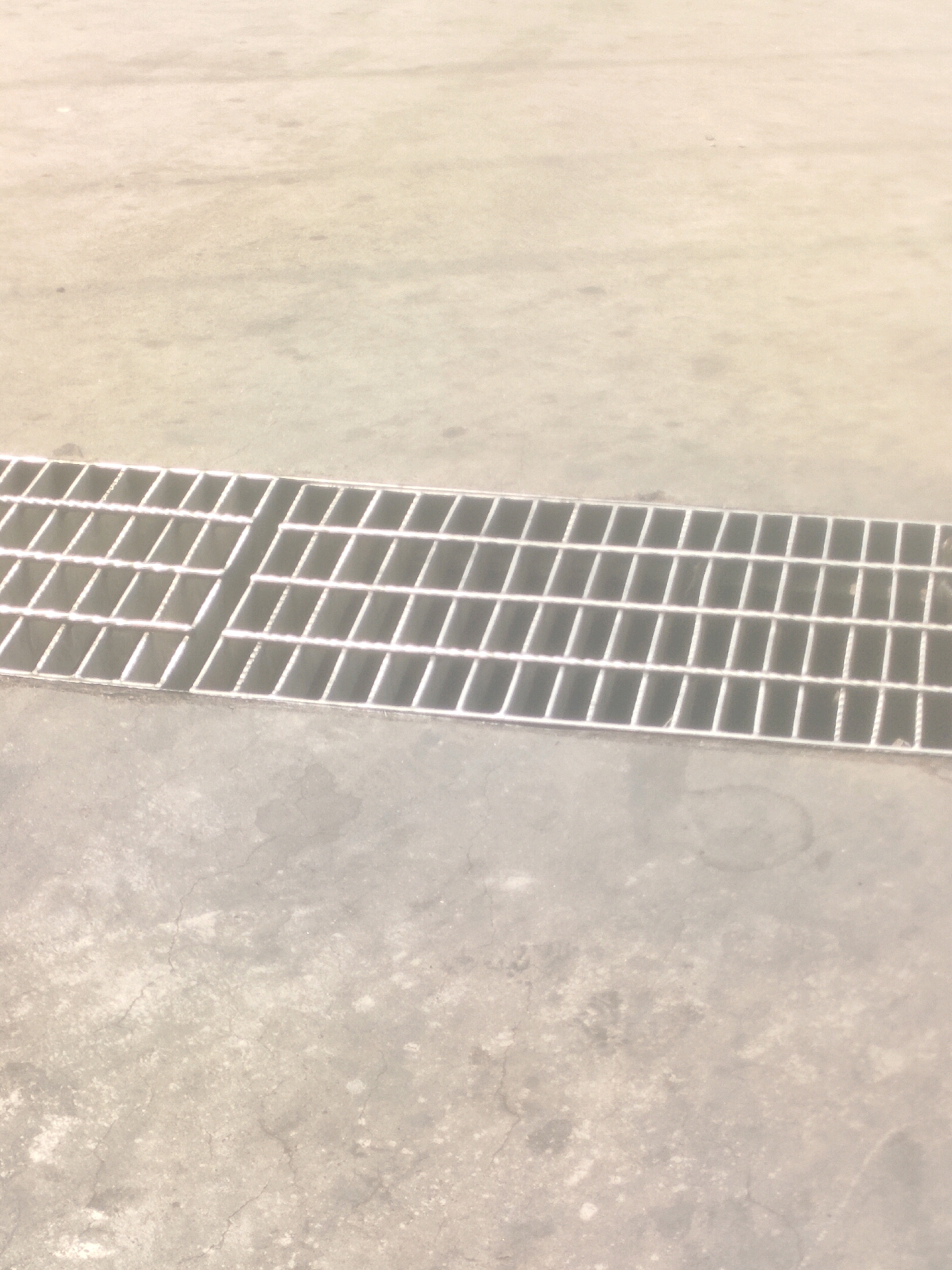 The image shows a close-up, high-angle view of a metal grate over a concrete drainage channel. The grate is made of parallel metal bars, forming a grid pattern. The concrete surface appears weathered and slightly stained. The lighting suggests an outdoor environment, possibly during the day, with diffuse light. There are no people or animals visible in the image. The scene is likely a public outdoor area in Surabaya, Indonesia, possibly a street, sidewalk, or parking lot.