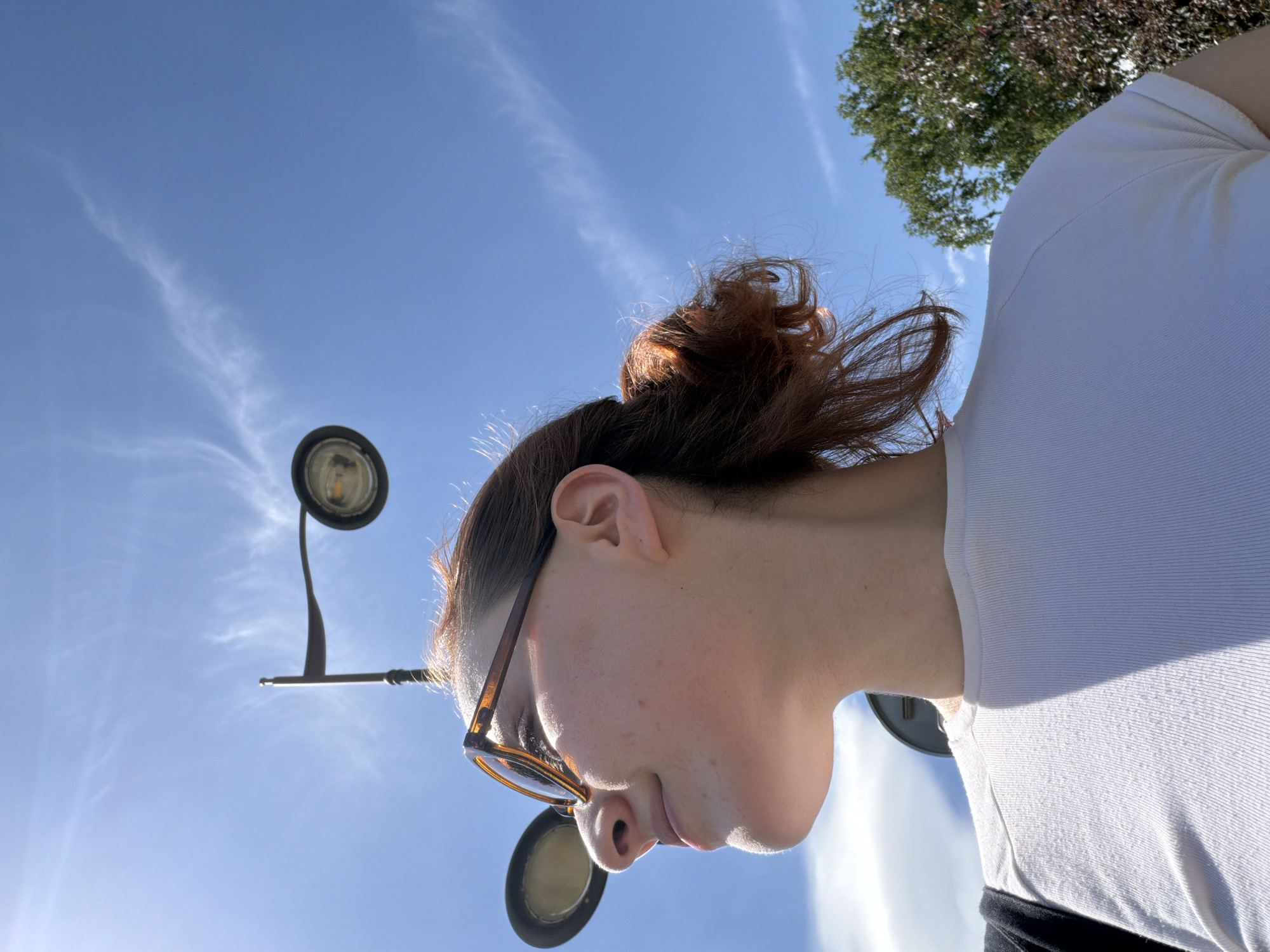 This is a low-angle shot of a young person with reddish-brown hair pulled into a bun, wearing sunglasses with tortoise-shell frames and a white t-shirt. They are looking downwards, and their face is illuminated by bright sunlight. The background is a clear blue sky with wispy white clouds. A tree is visible in the upper right corner. A dark, round object, possibly a lamp or a light fixture, is positioned to the left of the person's head, attached to a pole that extends into the frame. The overall impression is of a bright, sunny day, likely outdoors. The image is oriented sideways.