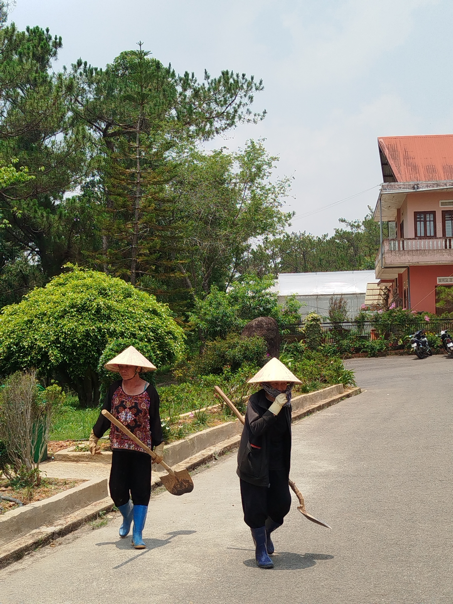 Two women in conical hats walk down an asphalt road in Lang Biang - Đà Lạt, Vietnam. The woman on the left wears a patterned top, black pants, and blue boots, and carries a shovel over her shoulder. The woman on the right wears a black jacket and pants, blue boots, and a scarf covering part of her face, and also carries a tool, possibly a hoe. The background includes trees, a building with a red roof, and parked motorcycles. A greenhouse structure is also visible. The sky is overcast.