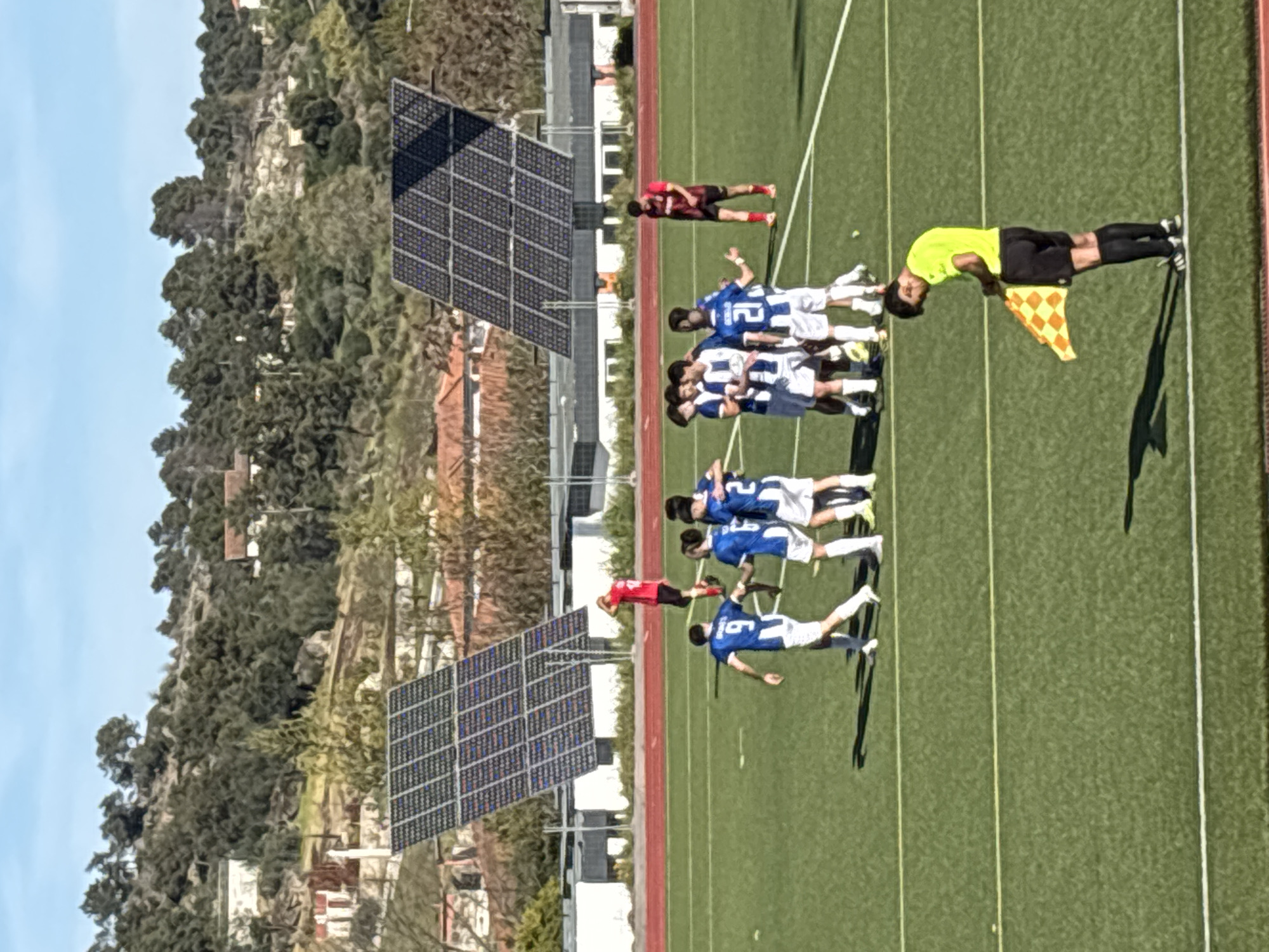 A soccer game is in progress on a green field in Valpaços, Portugal. A team wearing blue and white jerseys is grouped together, while a player in a red jersey lies on the ground nearby. A linesman in a yellow shirt and black shorts stands on the sideline, holding a yellow and orange checkered flag. In the background are two sets of solar panels, a red running track, a view of the city with houses and trees, and a blue sky.
