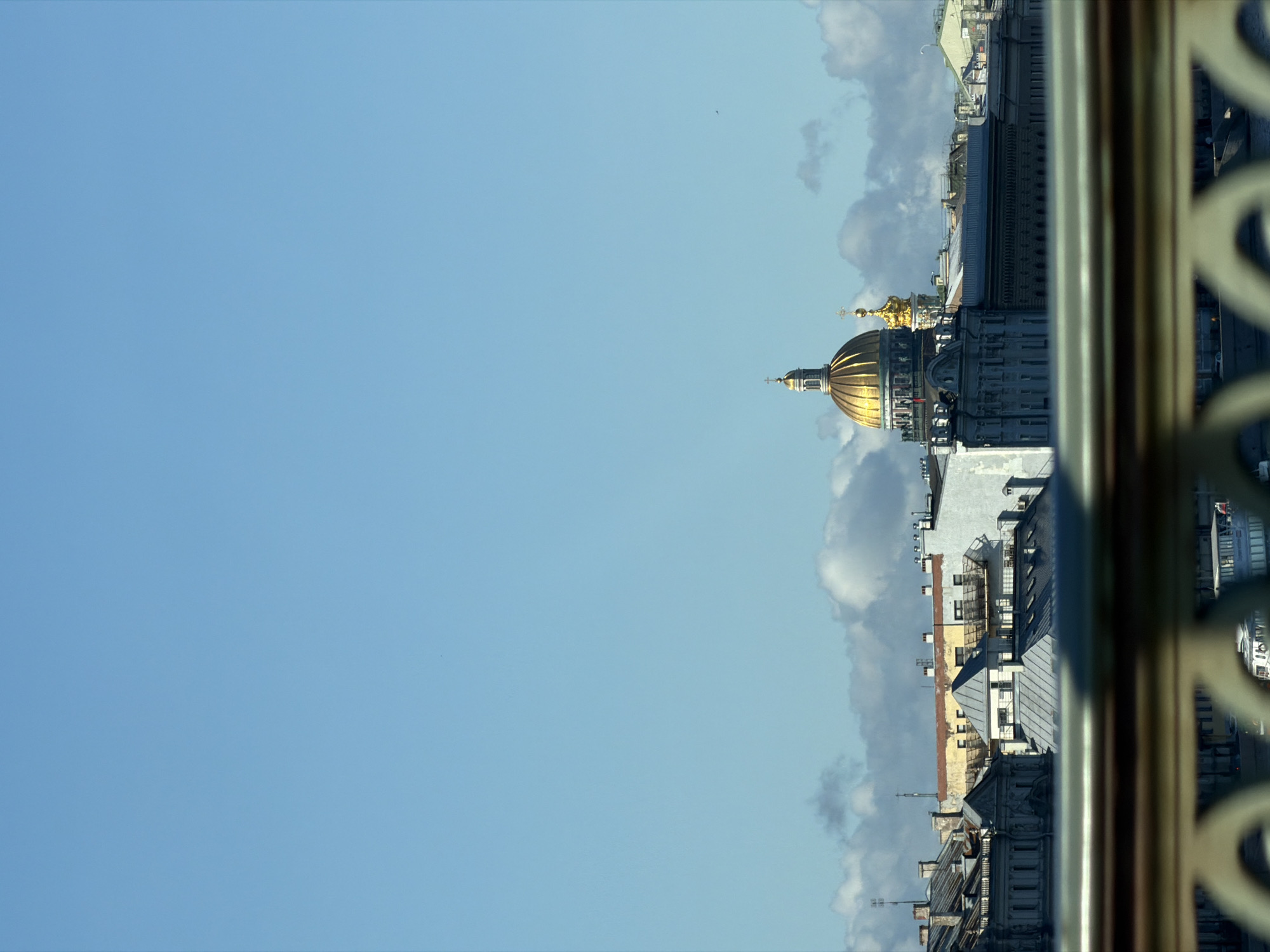 The image captures a cityscape in Saint Petersburg, Russia, dominated by a large, golden-domed building, likely a cathedral, with a prominent cross atop its spire. The architecture suggests a historical European city, with classical building styles evident in the surrounding structures. The sky is a clear, bright blue with scattered white clouds, indicating fair weather. In the foreground, a blurred railing with decorative elements suggests the viewer is on a balcony or bridge overlooking the scene. Below the main architectural subjects, a body of water is visible, with the outline of boats or ferries suggesting urban activity. The overall impression is of a grand, historic urban panorama under a clear sky.