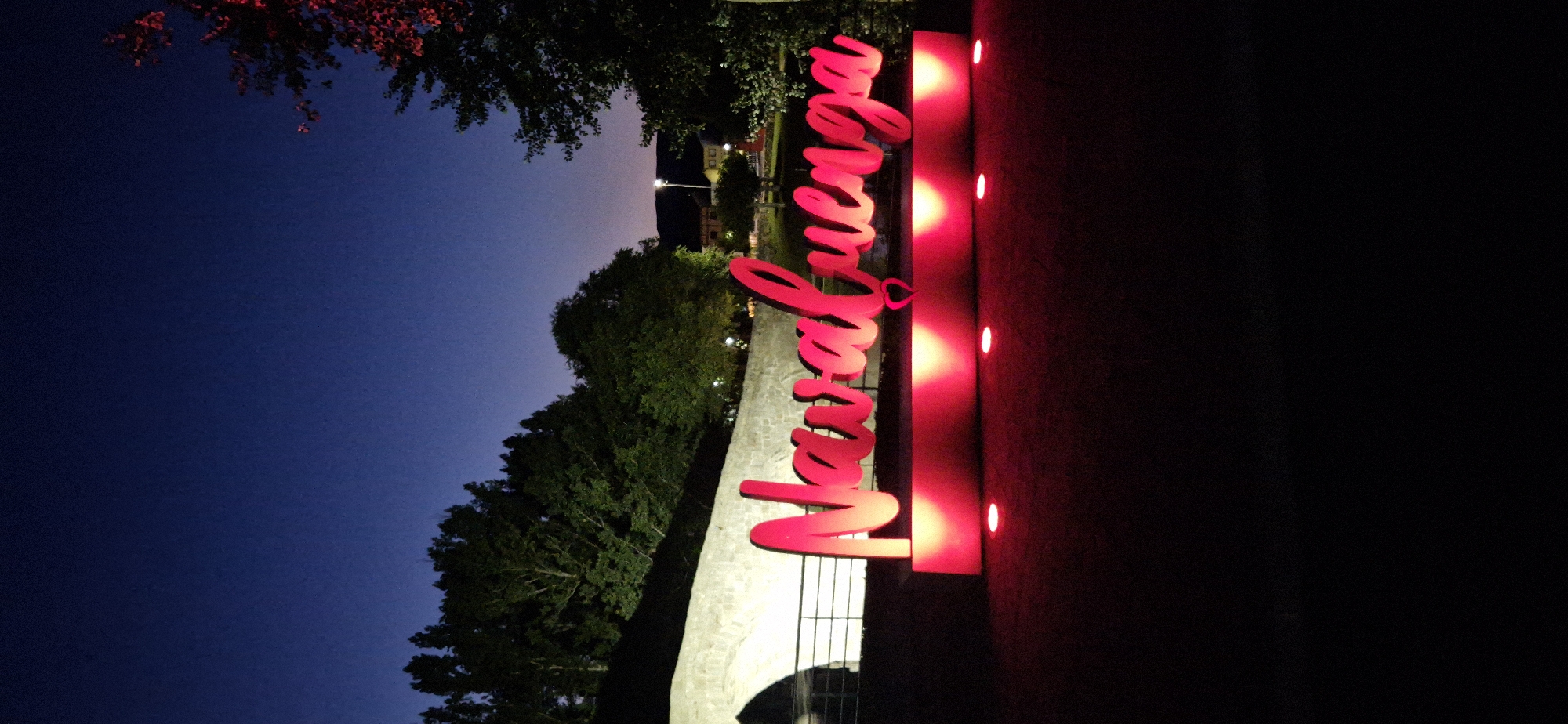 The image shows a night scene in Navaluenga, Spain, featuring a large, illuminated sign with the town's name in bright pink script. The sign is mounted on a red base that is also lit from below by a series of pink spotlights. The background includes a stone bridge and trees. The sky is a deep blue, suggesting it is twilight or nighttime. There are no people or animals visible in the image. The overall atmosphere is festive and inviting.