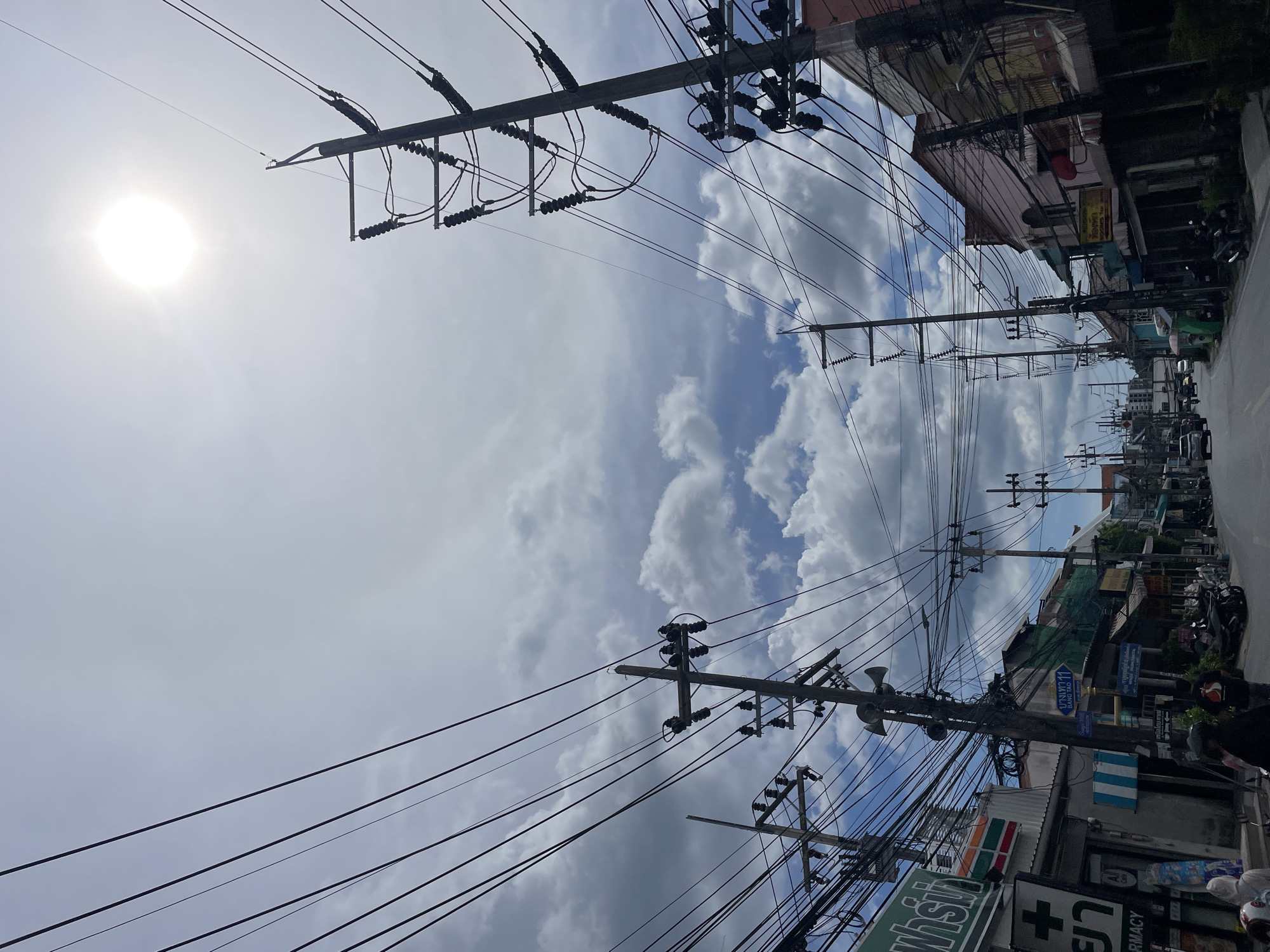 An upward-angled, medium shot depicts a street in Choeng Thale, Thailand, under a bright sky with scattered clouds. Numerous overhead power lines dominate the view. Several utility poles stretch along the street, loaded with transformers and electrical equipment. Buildings line both sides of the street, with visible signage including a 7-Eleven logo and other Thai script signs. Parked motorcycles are visible along the right side of the street. The sun is visible in the upper left portion of the image. A pharmacy sign is visible on the lower right. The street appears to stretch into the distance.