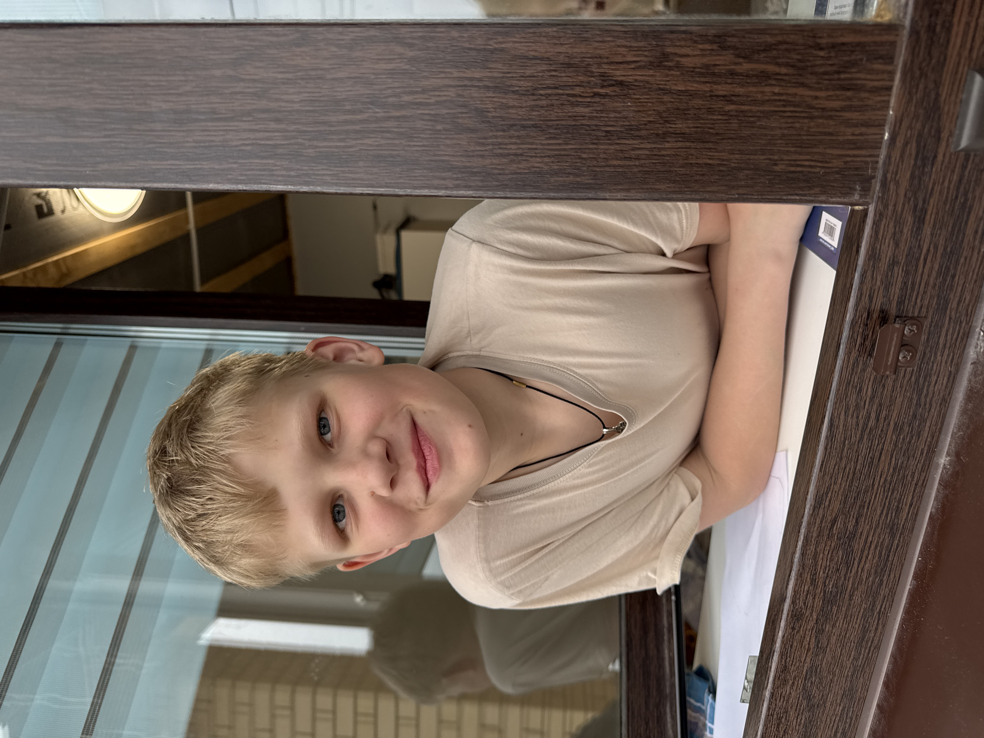The image shows a young male individual, estimated to be a child or adolescent, positioned partially through an open brown-framed window in Gornyi (Dvurechenskoe S/P), Russia. The individual has blond hair, blue eyes, and fair skin, and is wearing a light tan short-sleeved t-shirt and a dark necklace with a light-colored pendant. He is leaning forward, resting his forearms on a white surface, possibly a windowsill or desk, which holds papers and a blue object with a visible barcode. He is looking directly at the viewer with a slight smile.
The window frame is dark brown with a wood-grain texture. Through the open section of the window to the right of the individual, an interior space is visible, featuring an inclined ceiling with exposed wooden beams and an illuminated circular light fixture. The window glass on the left reflects an exterior brick wall, appearing light-colored, and a blurry reflection resembling another person. The bottom section of the window frame shows some dust accumulation.